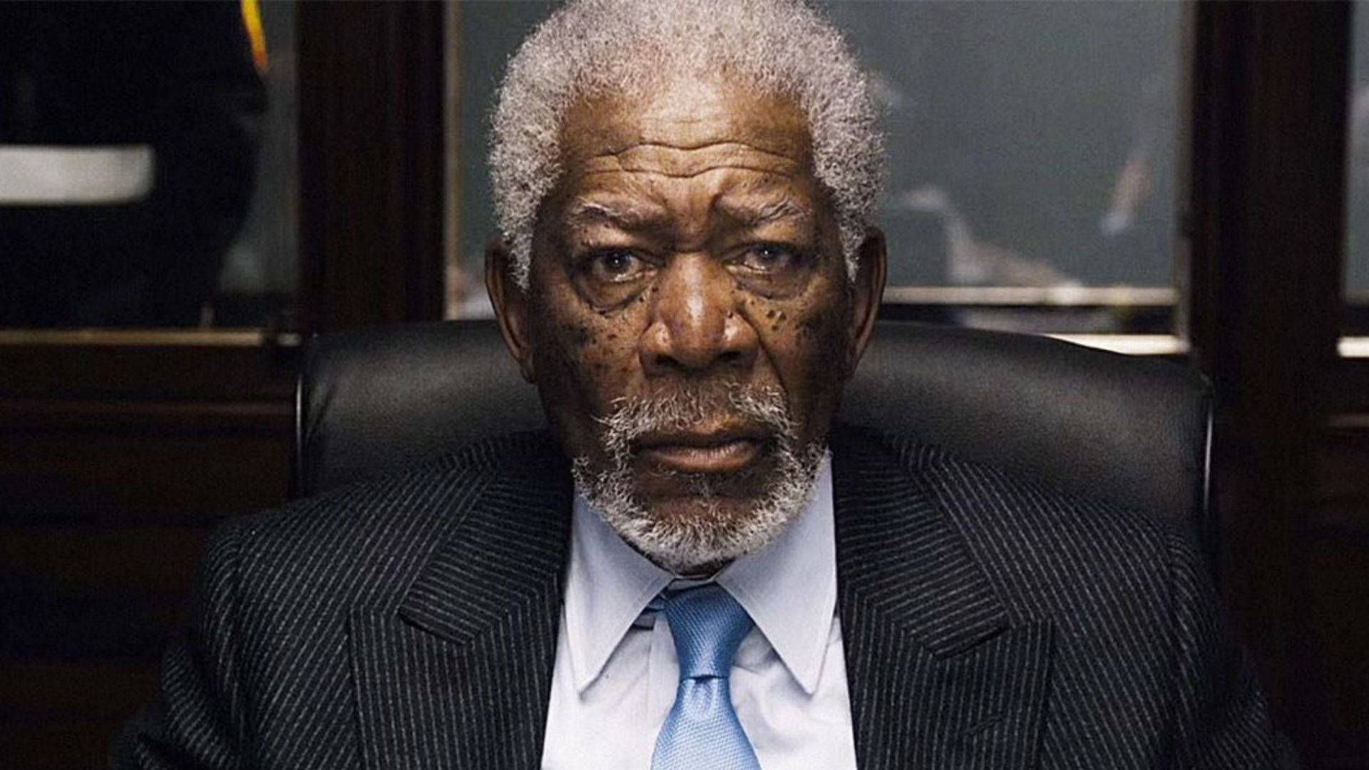 Morgan Freeman 桌布 壁紙 — 一位年長的黑人男性，灰白頭髮和鬍鬚，坐在黑色皮椅上，穿著細條紋西裝和藍色領帶，專注地向前看。