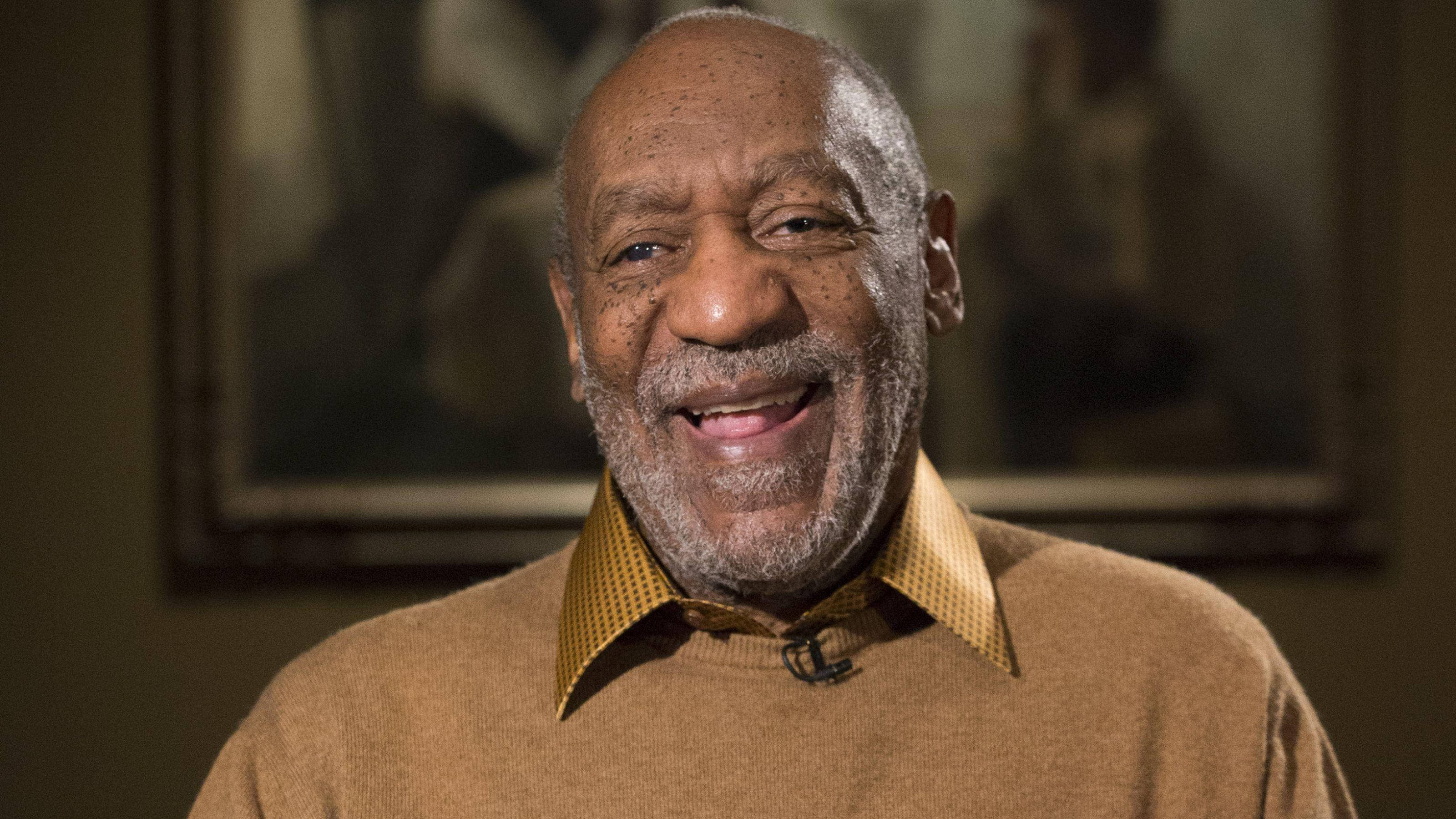 bill cosby 桌布 壁紙 — 一位年長的黑人男性，面帶溫暖笑容，灰色鬍鬚，室內穿著米色毛衣內搭黃色圖案襯衫。
