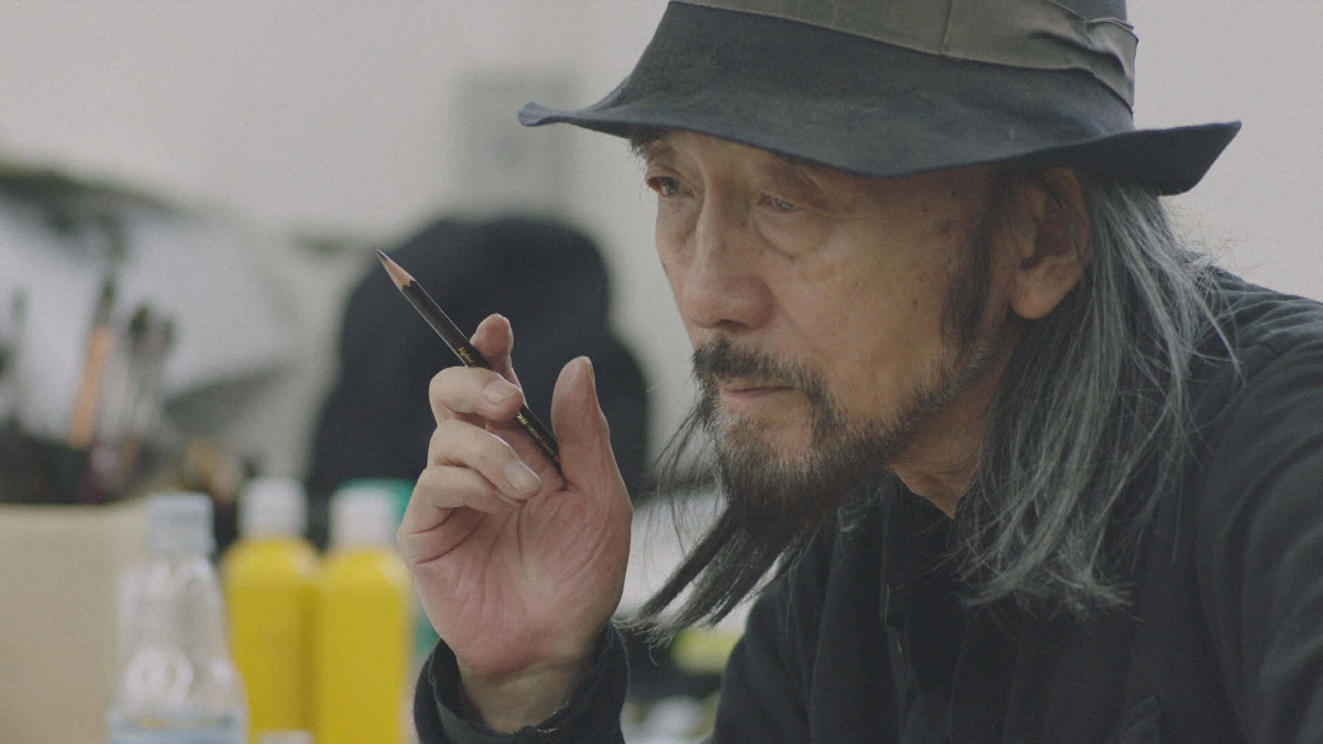 時尚 桌布 壁紙 — 一位年長藝術家，留著灰色長髮，戴著帽子，在充滿美術用品的畫室裡若有所思地拿著鉛筆。