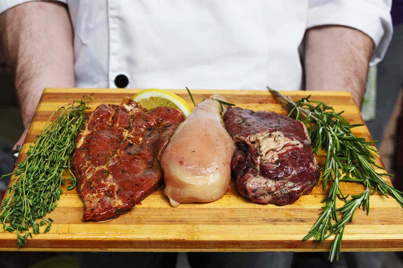 meat 桌布 壁紙 — 一位廚師展示一個木盤，上面有調味牛排、雞胸肉和羊肉，配以新鮮香草和檸檬裝飾。
