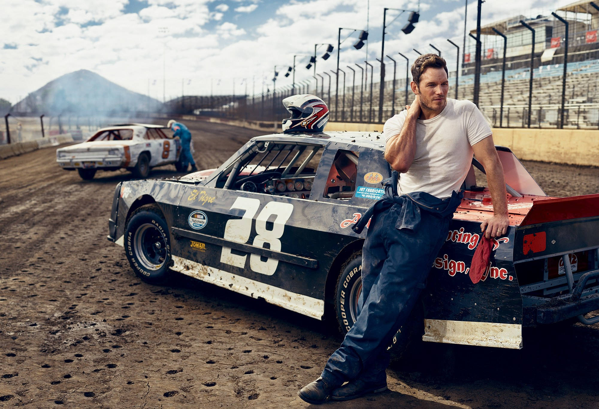 Chris Pratt 桌布 壁紙 — 一位強壯的賽車手在晴朗的天空下，在塵土飛揚的泥地賽道上，隨意地靠在他那濺滿泥漿的28號改裝車上。