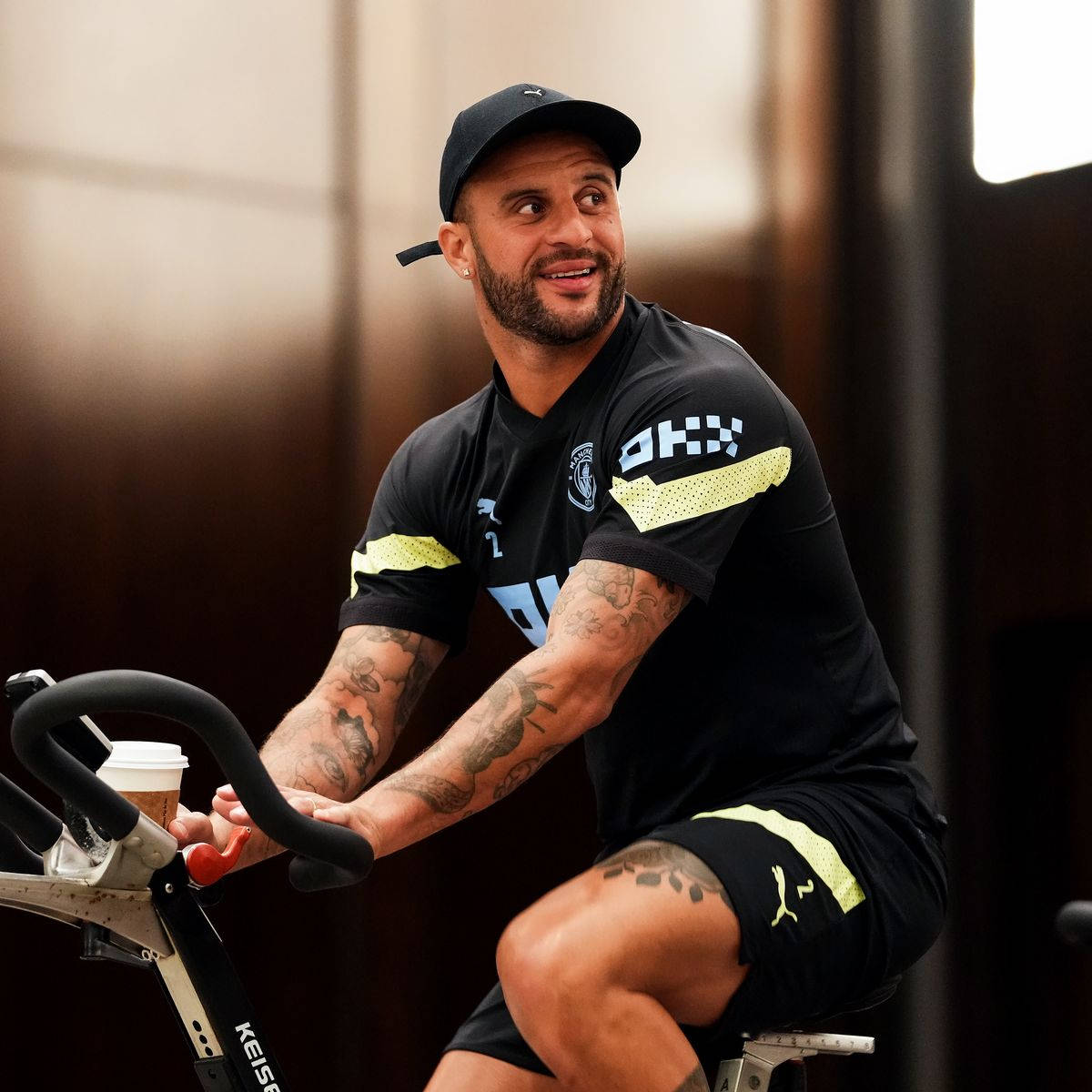 Kyle Walker 桌布 壁紙 — 一位微笑、有刺青的男士穿著黑色運動服，在室內騎固定式腳踏車，手持一杯咖啡。