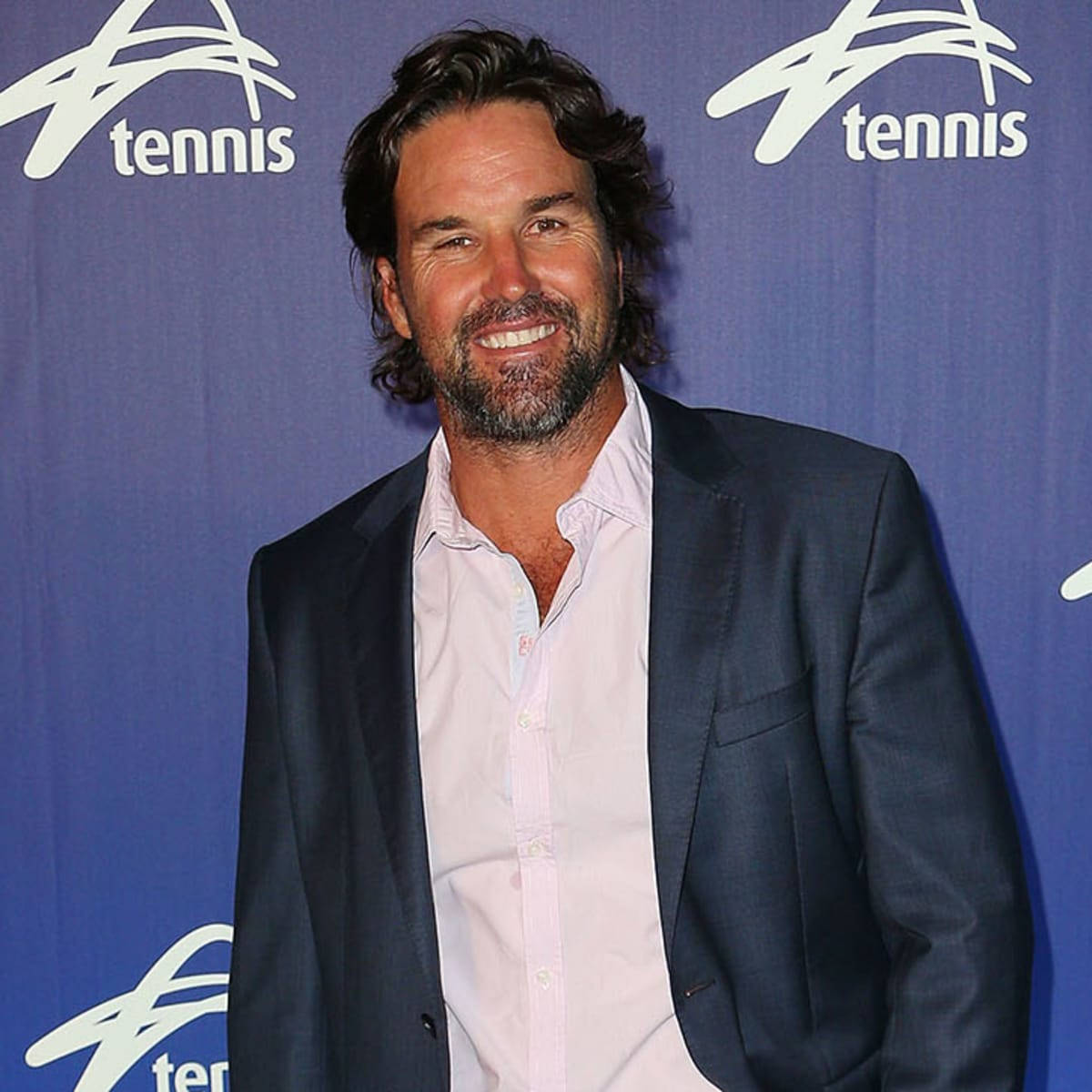 Tennis 桌布 壁紙 — 一位微笑、留鬍鬚、深色頭髮的男子，身穿海軍藍西裝外套，內搭淺粉紅色襯衫，背景為藍色網球品牌背景。