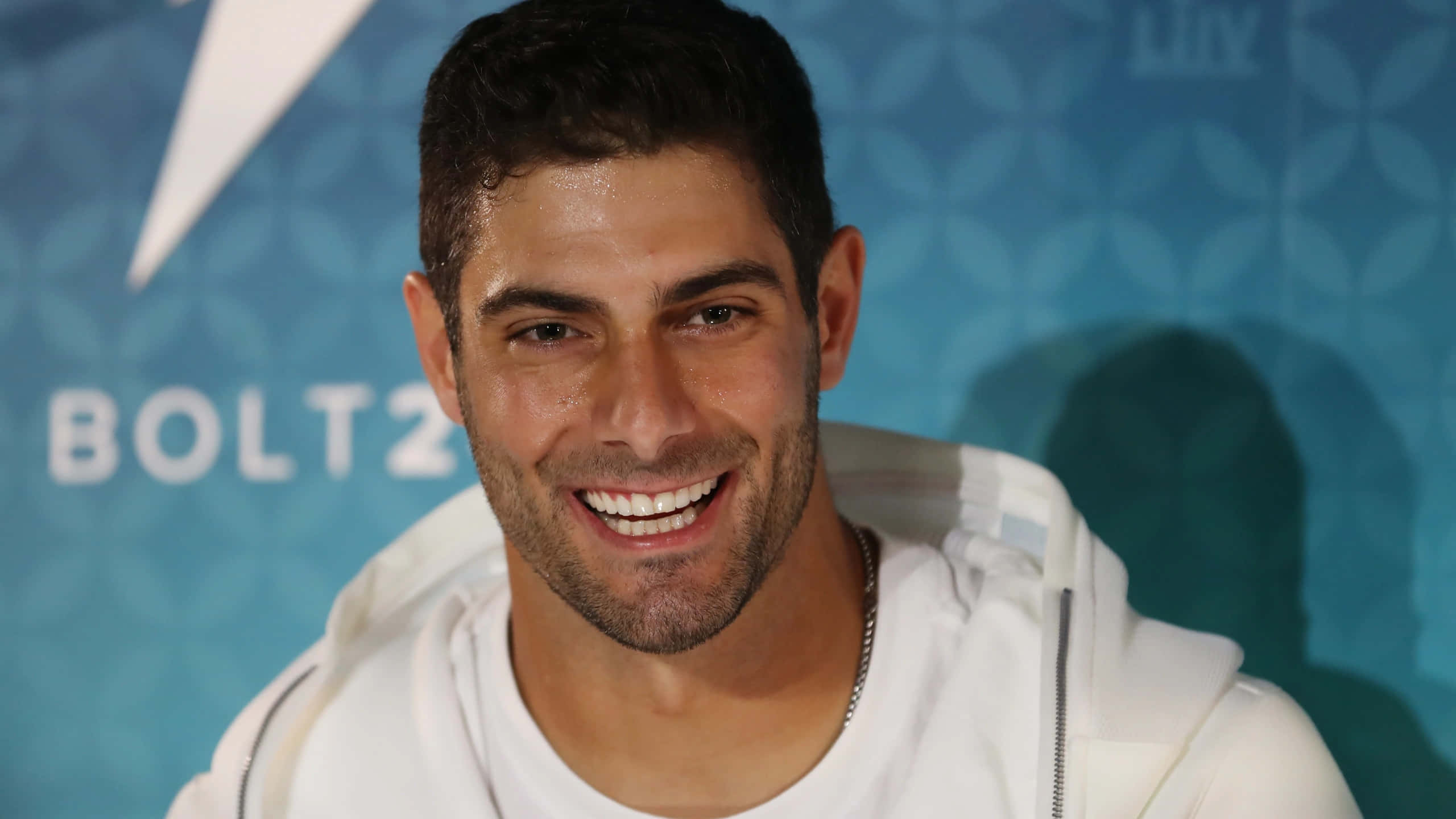 Jimmy Garoppolo 桌布 壁紙 — 一位微笑、短黑髮、鬍渣的男子，身穿白色連帽衫，背景為藍色圖案並帶有 'BOLT2' 文字。
