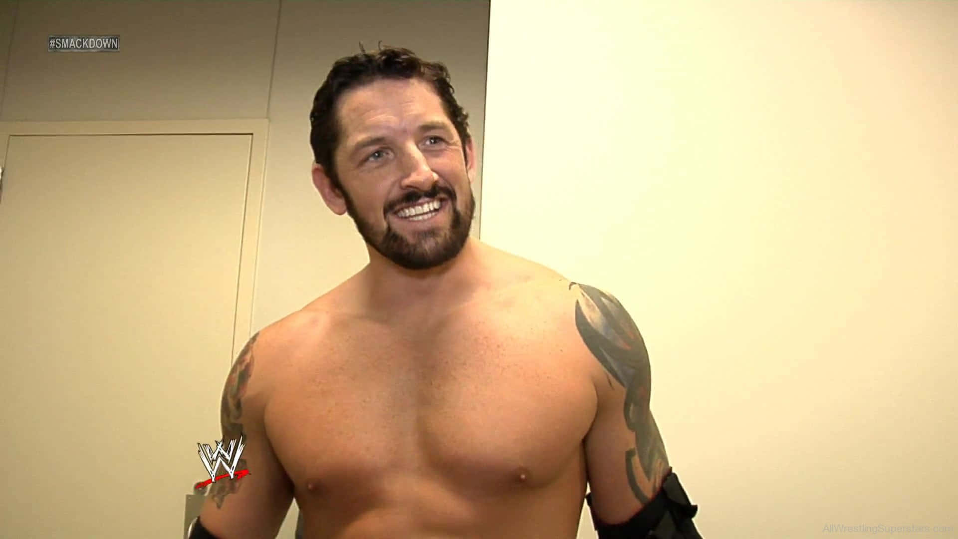 WWE 桌布 壁紙 — 一位微笑、肌肉發達、手臂上有刺青的WWE摔角手，赤裸上身站在後台走廊。