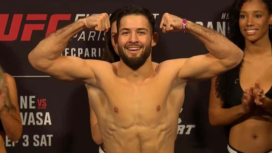 MMA 桌布 壁紙 — 一位微笑、肌肉發達的UFC拳手在稱重活動中彎曲手臂，背景顯示比賽詳情。