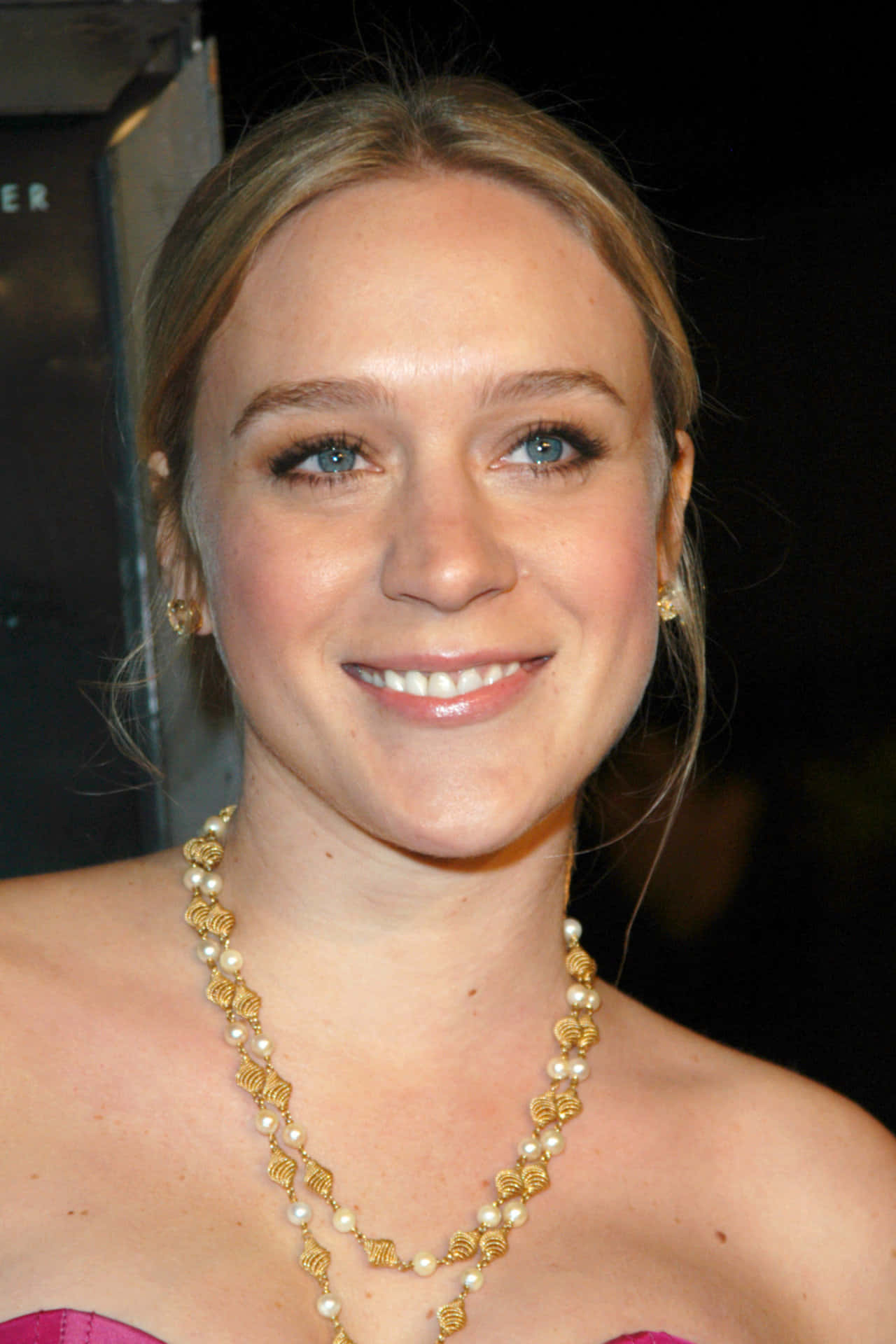Chloë Sevigny 桌布 壁紙 — 一位微笑女士的近距离肖像，藍色眼睛，戴著金和珍珠項鍊，穿著粉紅色連衣裙。