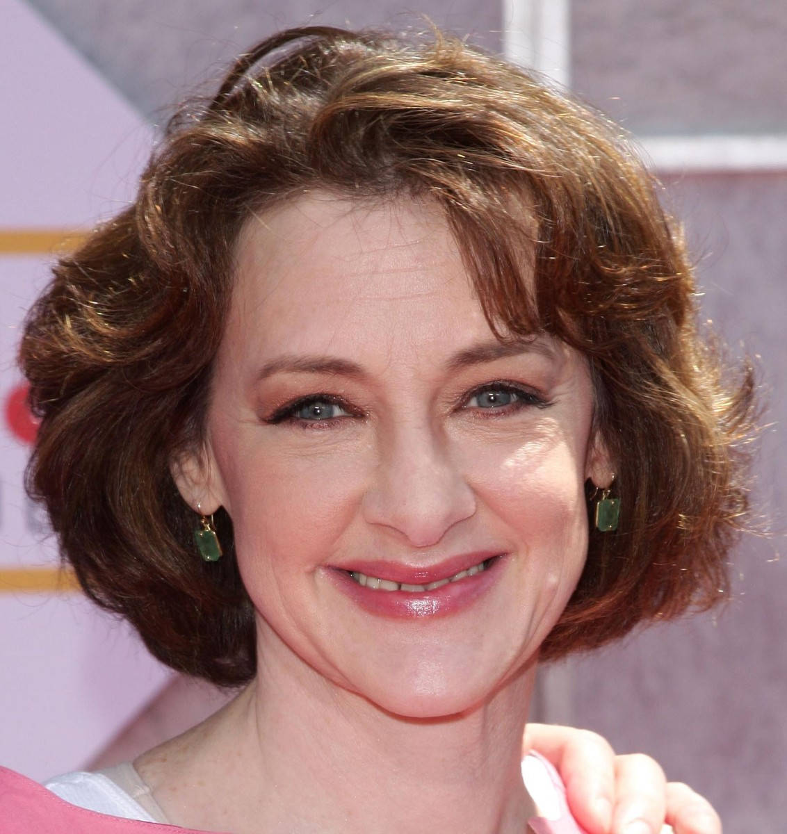 Celebrity 桌布 壁紙 — 一位微笑女士的頭像特寫，波浪棕色頭髮和綠色耳環，背景為柔和粉紅色。