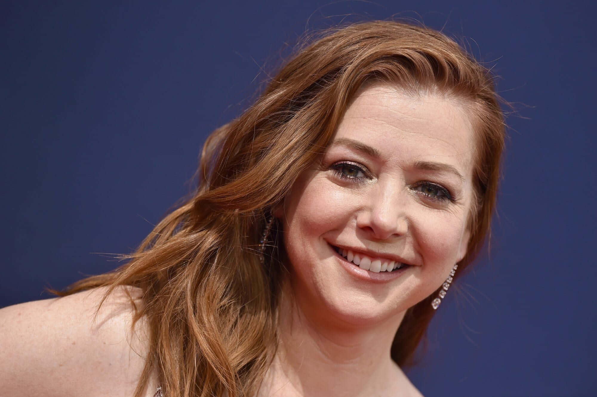 Alyson Hannigan 桌布 壁紙 — 一位微笑女士的頭像近照，擁有波浪形赤褐色頭髮，背景為純藍色。