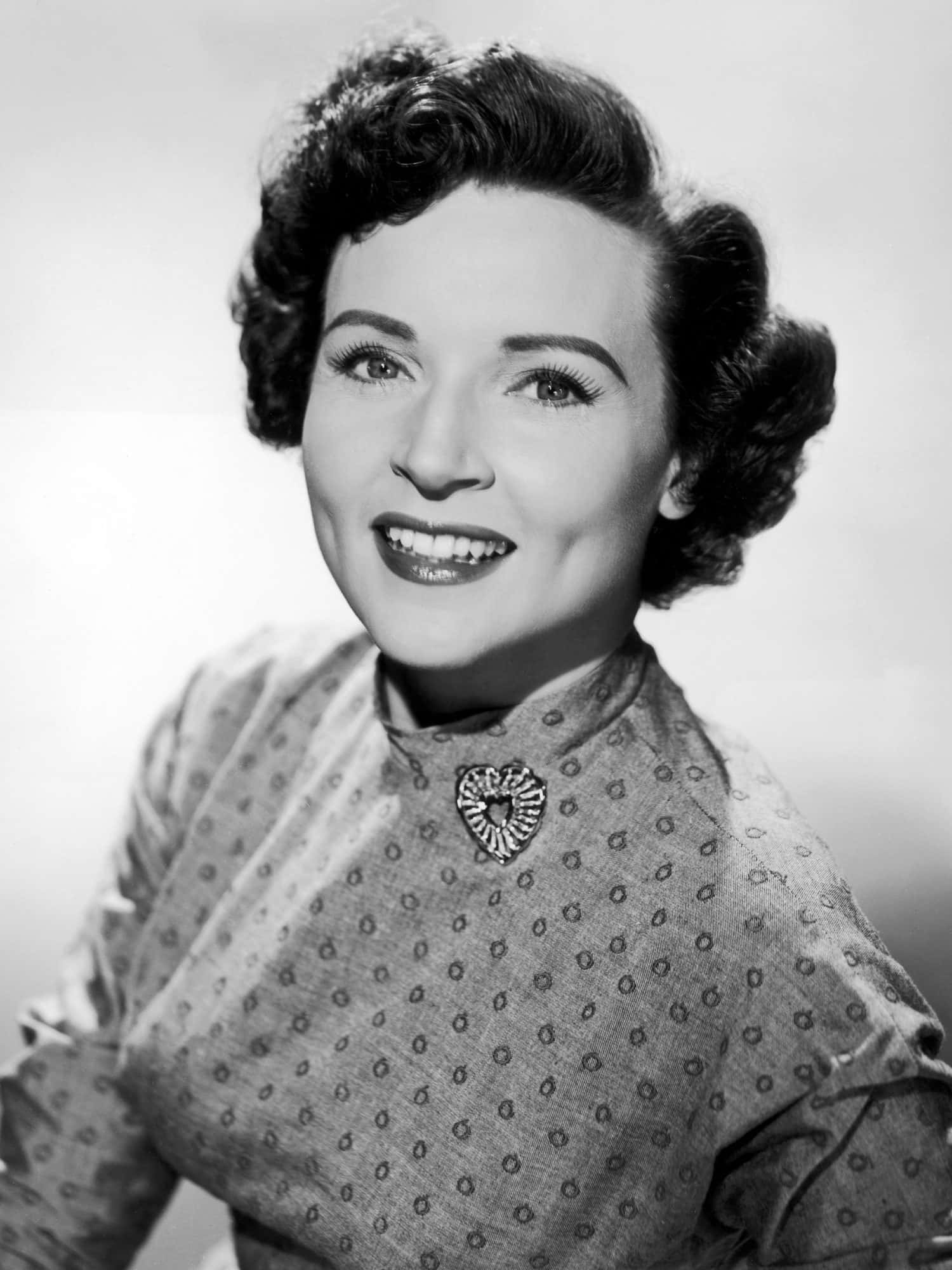 Betty White 桌布 壁紙 — 一位微笑女性的復古黑白肖像，她有造型捲髮和帶有心形胸針的圖案襯衫。