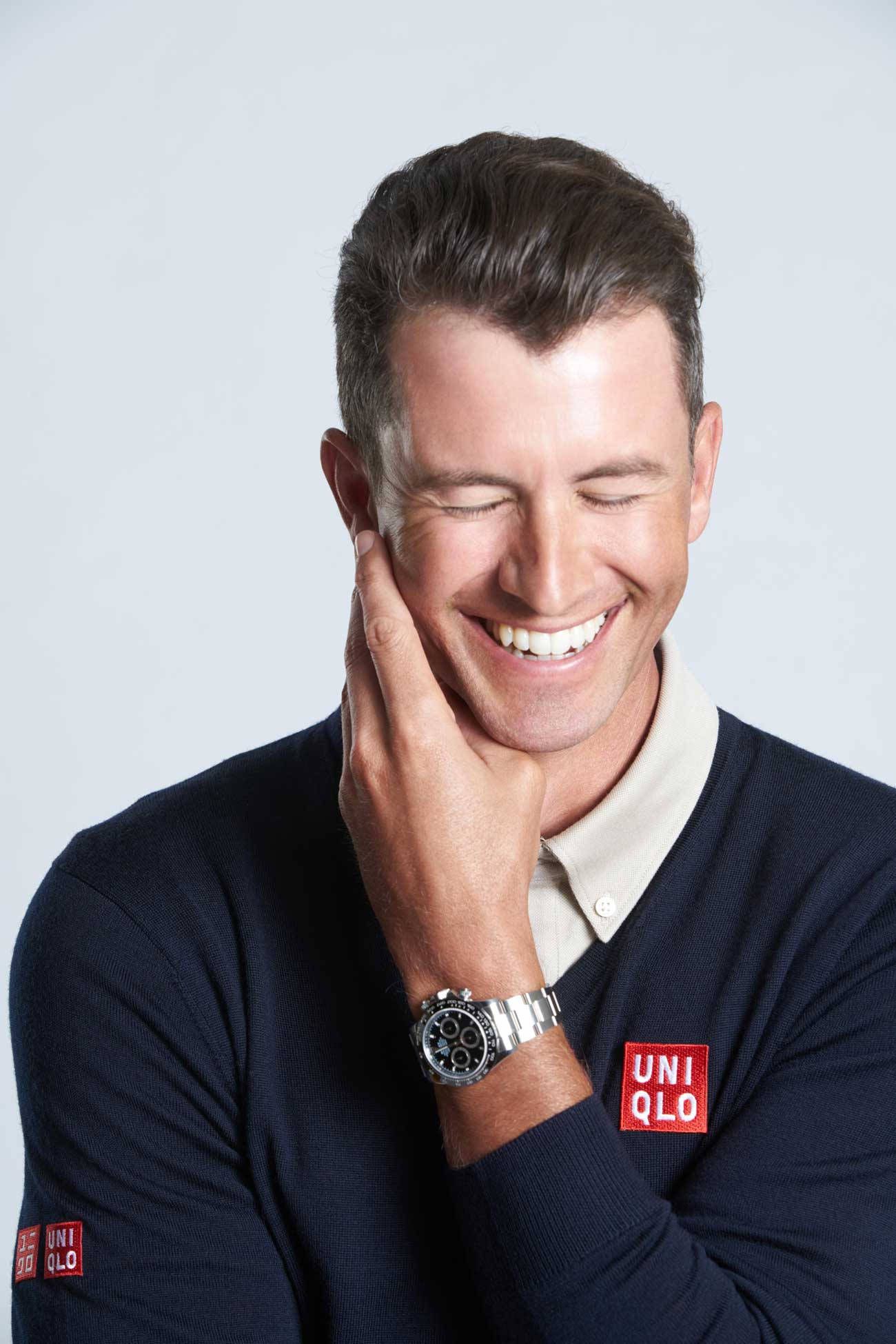 adam scott 桌布 壁紙 — 一位微笑男士手托臉頰，身穿深藍色Uniqlo毛衣及銀色手錶，背景簡潔。