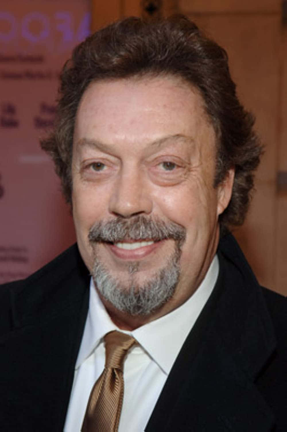 Tim Curry 桌布 壁紙 — 一位微笑男士的特寫肖像，留著灰白鬍鬚，身穿黑色西裝和金色領帶。