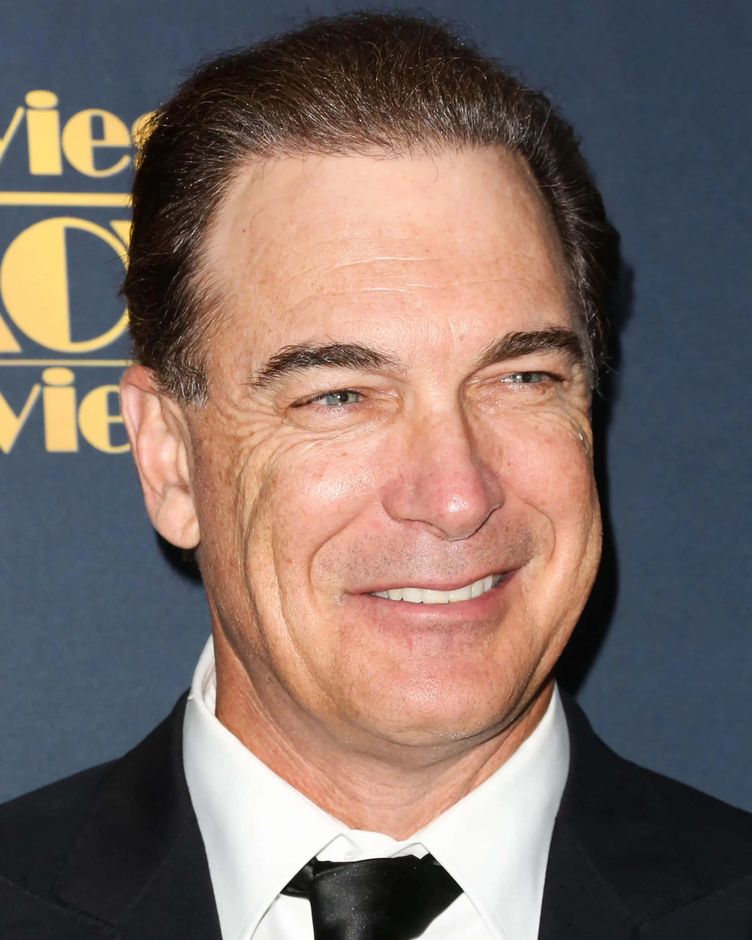 Patrick Warburton 桌布 壁紙 — 一位微笑男士的近距離肖像，身穿黑色西裝和領帶，背景為藍色並帶有金色字樣。