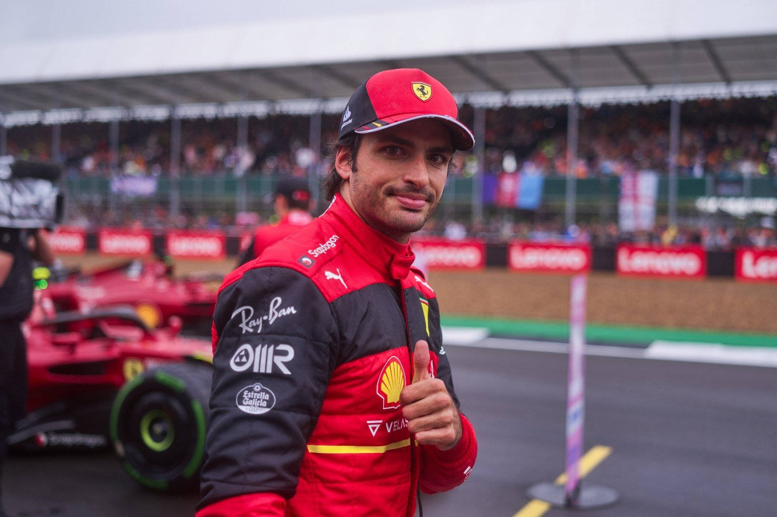 Racing 桌布 壁紙 — 一位微笑的F1車手穿著紅色法拉利裝備，在賽道上他的賽車旁邊豎起大拇指，背景有人群。