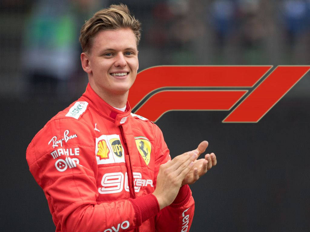 mick schumacher 桌布 壁紙 — 一位微笑的F1車手身穿紅色法拉利賽車服，在大型F1標誌前拍手。