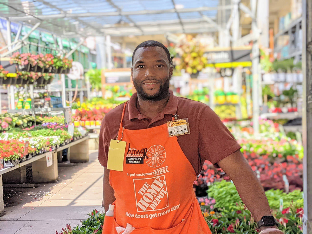 Home Depot 桌布 壁紙 — 一位微笑的Home Depot助理站在明亮的溫室通道中，周圍環繞著色彩繽紛的盆栽花卉和植物。
