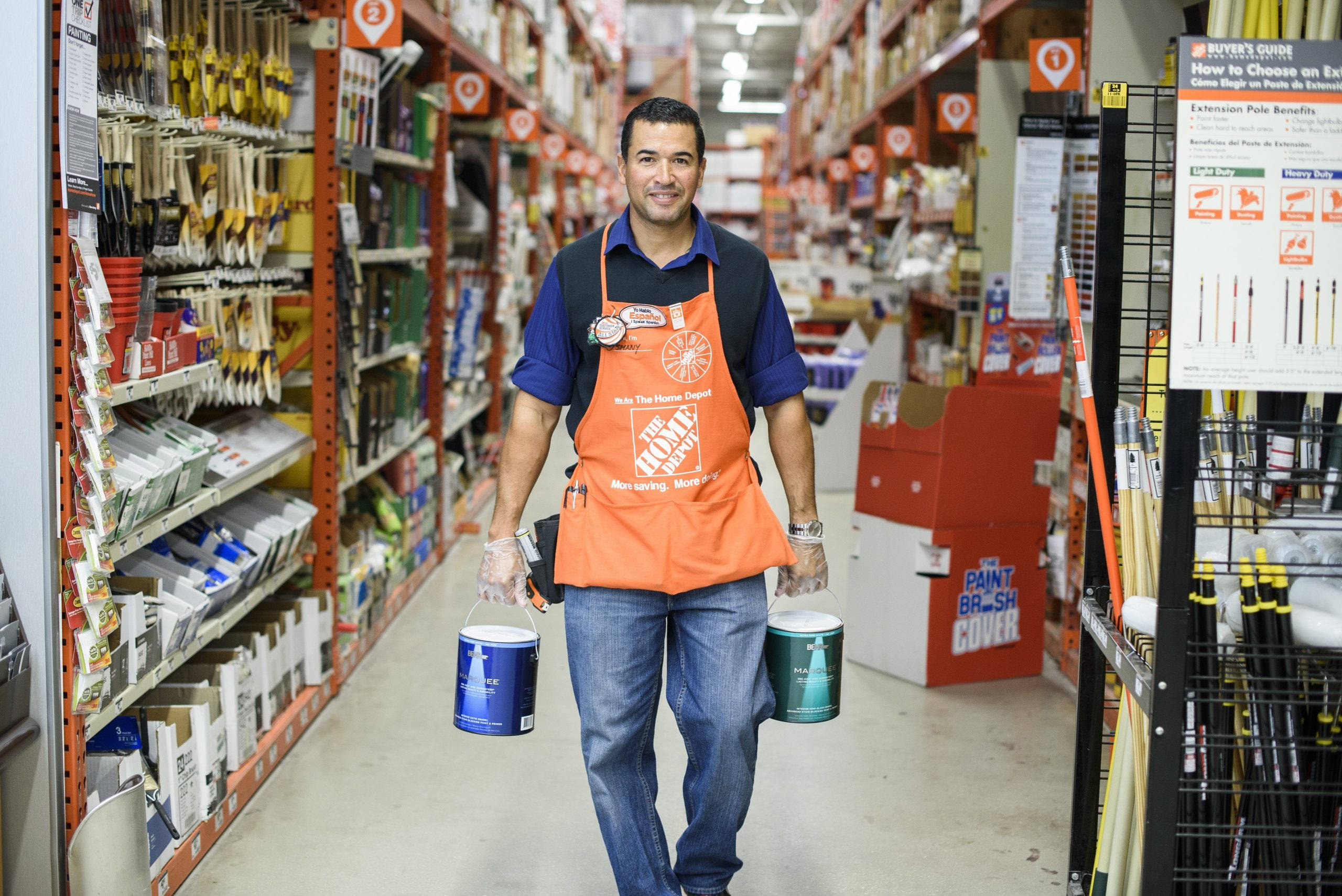 Home Depot 桌布 壁紙 — 一位微笑的Home Depot員工穿著橙色圍裙，在光線充足的五金店走廊上搬運兩罐油漆，走廊兩旁擺滿工具和用品。