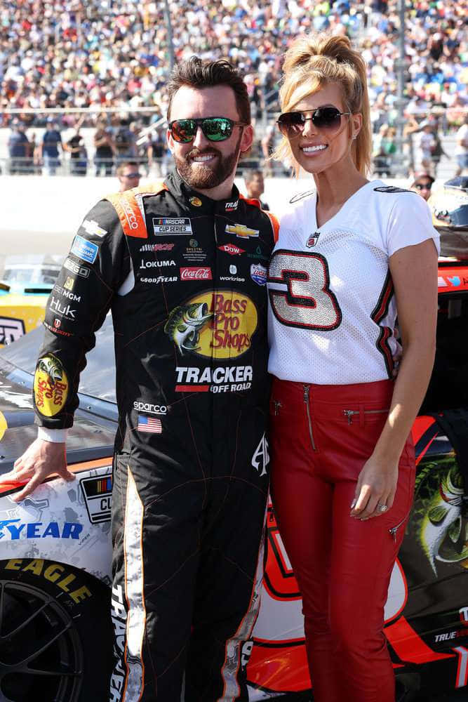 nascar 桌布 壁紙 — 一位微笑的NASCAR車手和一位金髮賽車女郎在陽光明媚的賽道上，於一輛賽車旁擺姿勢，賽道擠滿了觀眾。