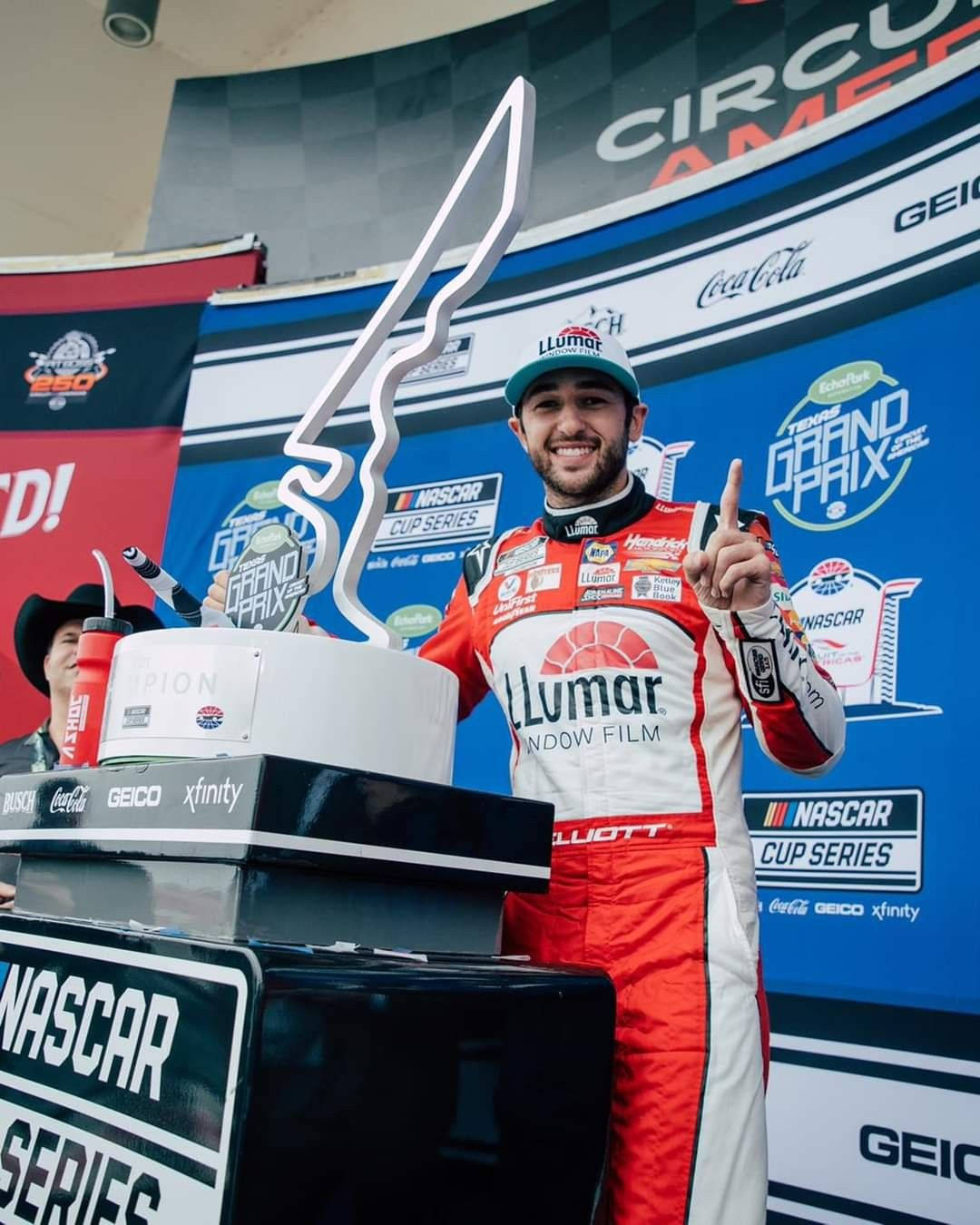 Chase Elliott 桌布 壁紙 — 一位微笑的NASCAR車手身穿紅白賽車服，站在頒獎台上，手持大獎盃並向上指，在德州大獎賽活動中。