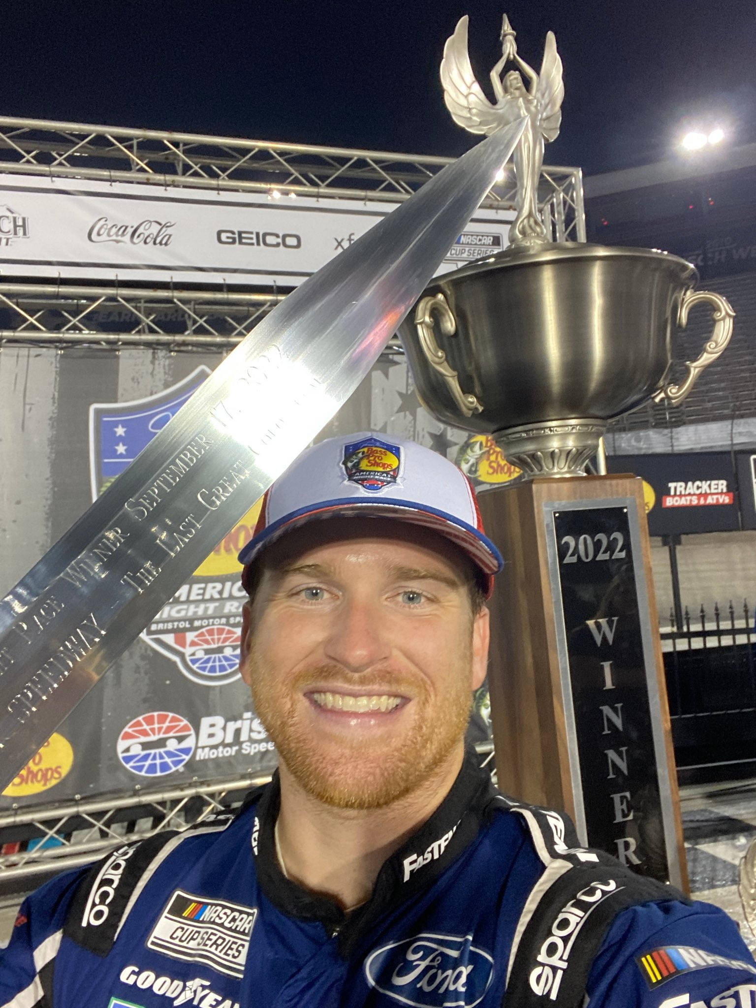 Champion 桌布 壁紙 — 一位微笑的NASCAR車手身穿藍色賽車服，在夜晚自豪地舉起一個巨大的銀色獎盃。