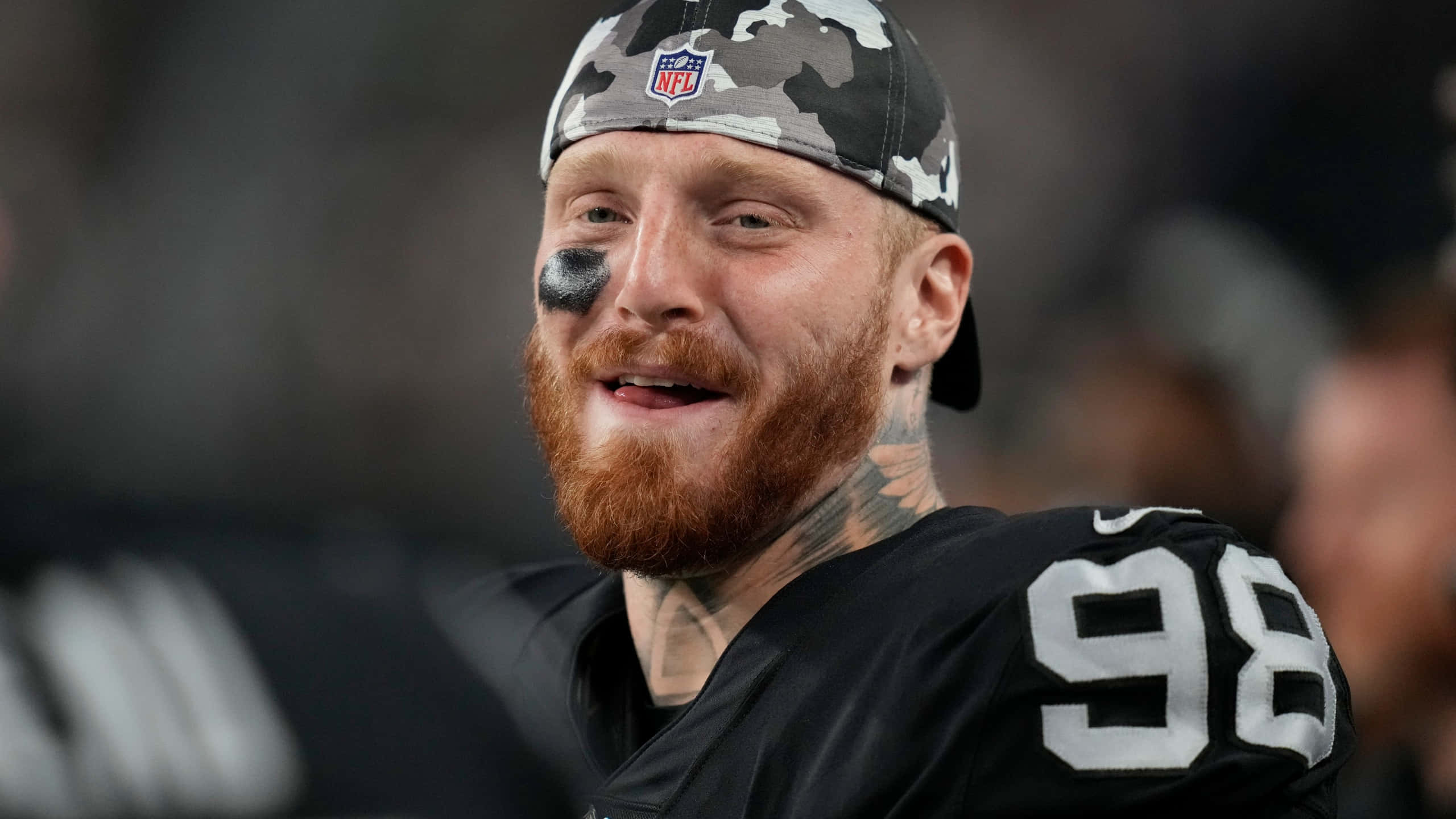 Las Vegas Raiders 桌布 壁紙 — 一位微笑的NFL球員，留著紅色鬍鬚，眼睛塗有黑色顏料，戴著迷彩帽，身穿98號球衣。