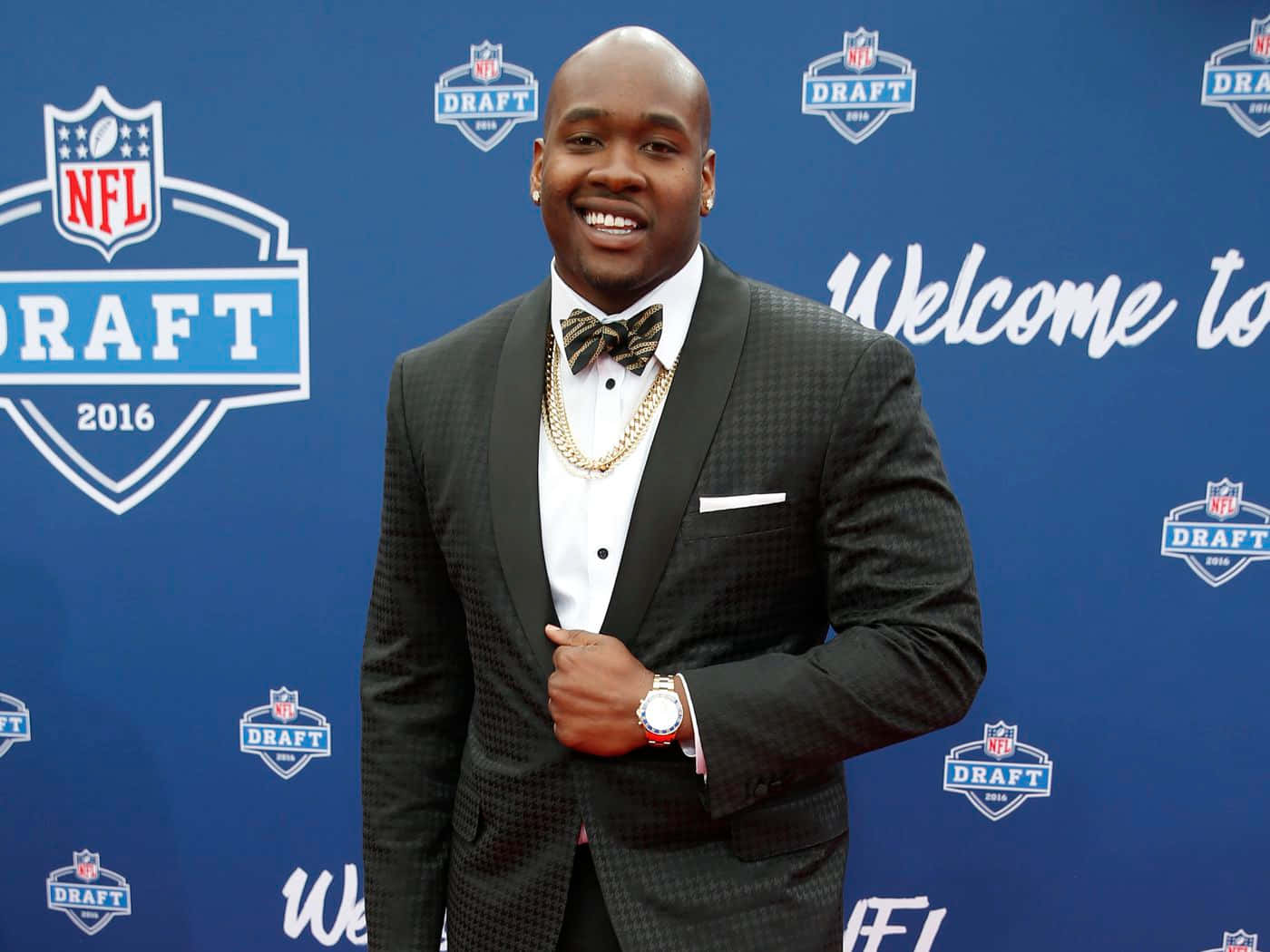 足球 桌布 壁紙 — 一位微笑的NFL選秀新秀穿著圖案禮服和金色鏈條，在藍色活動背景前自信地擺姿勢。