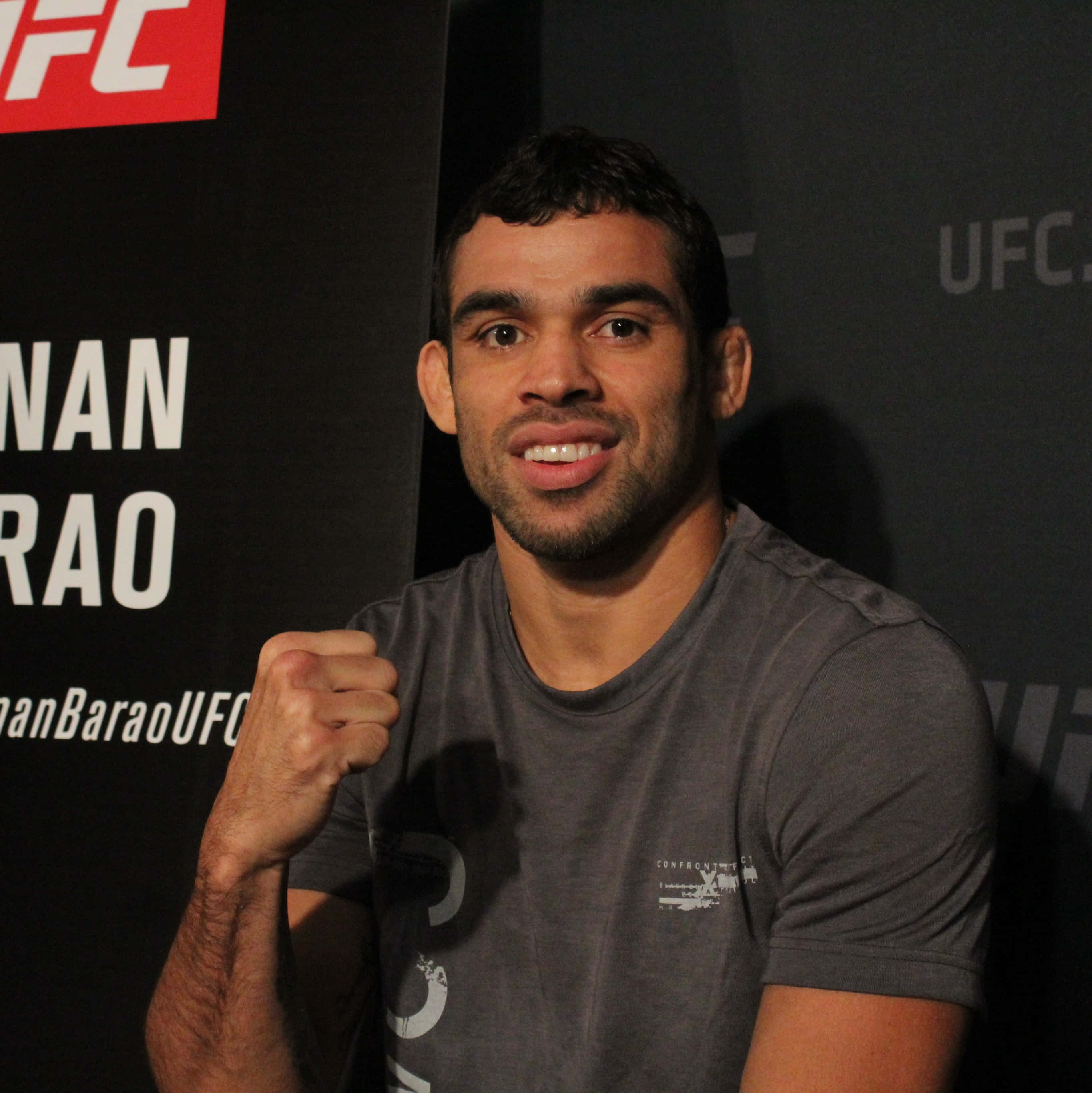UFC 桌布 壁紙 — 一位微笑的UFC拳手身穿灰色T恤，在品牌背景前握拳擺姿勢。