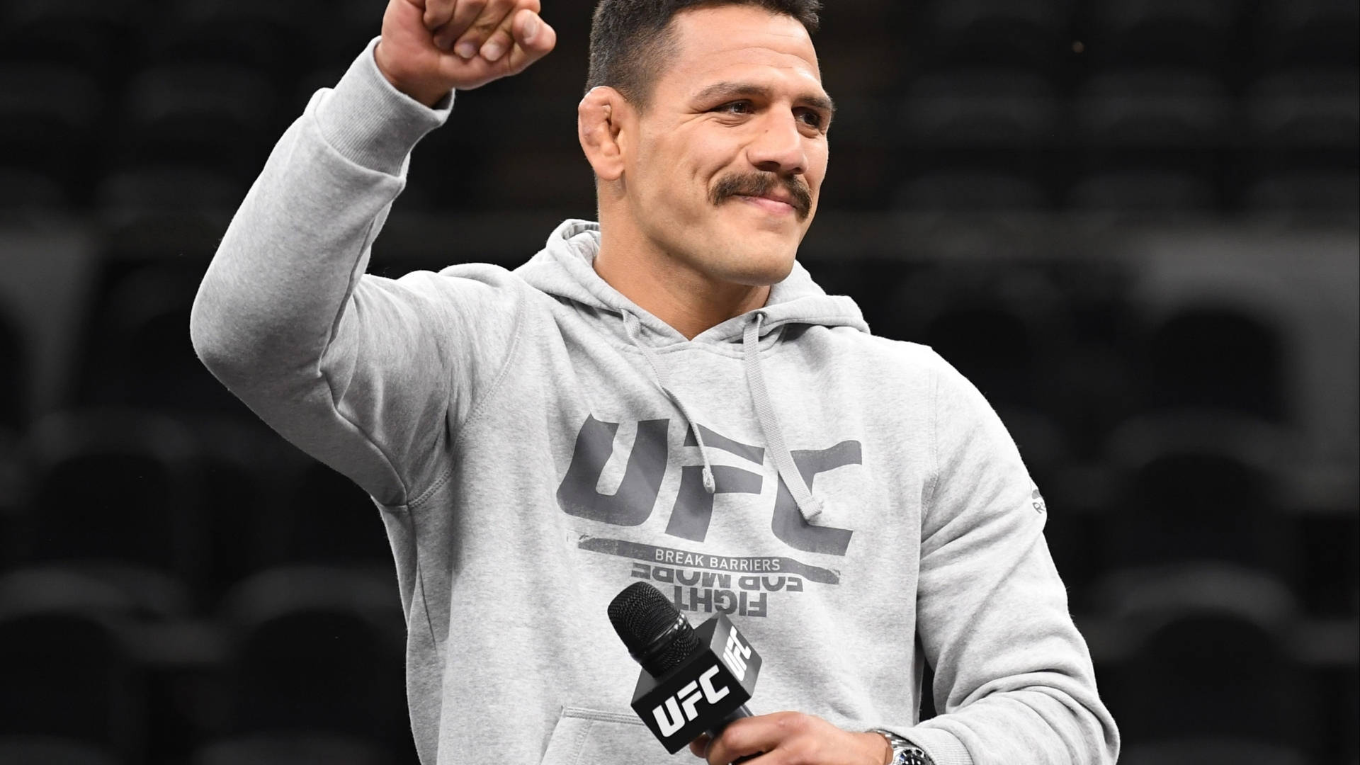 UFC 桌布 壁紙 — 一位微笑的UFC拳手身穿灰色連帽衫，在燈光昏暗的場館中舉起拳頭，同時手持麥克風。