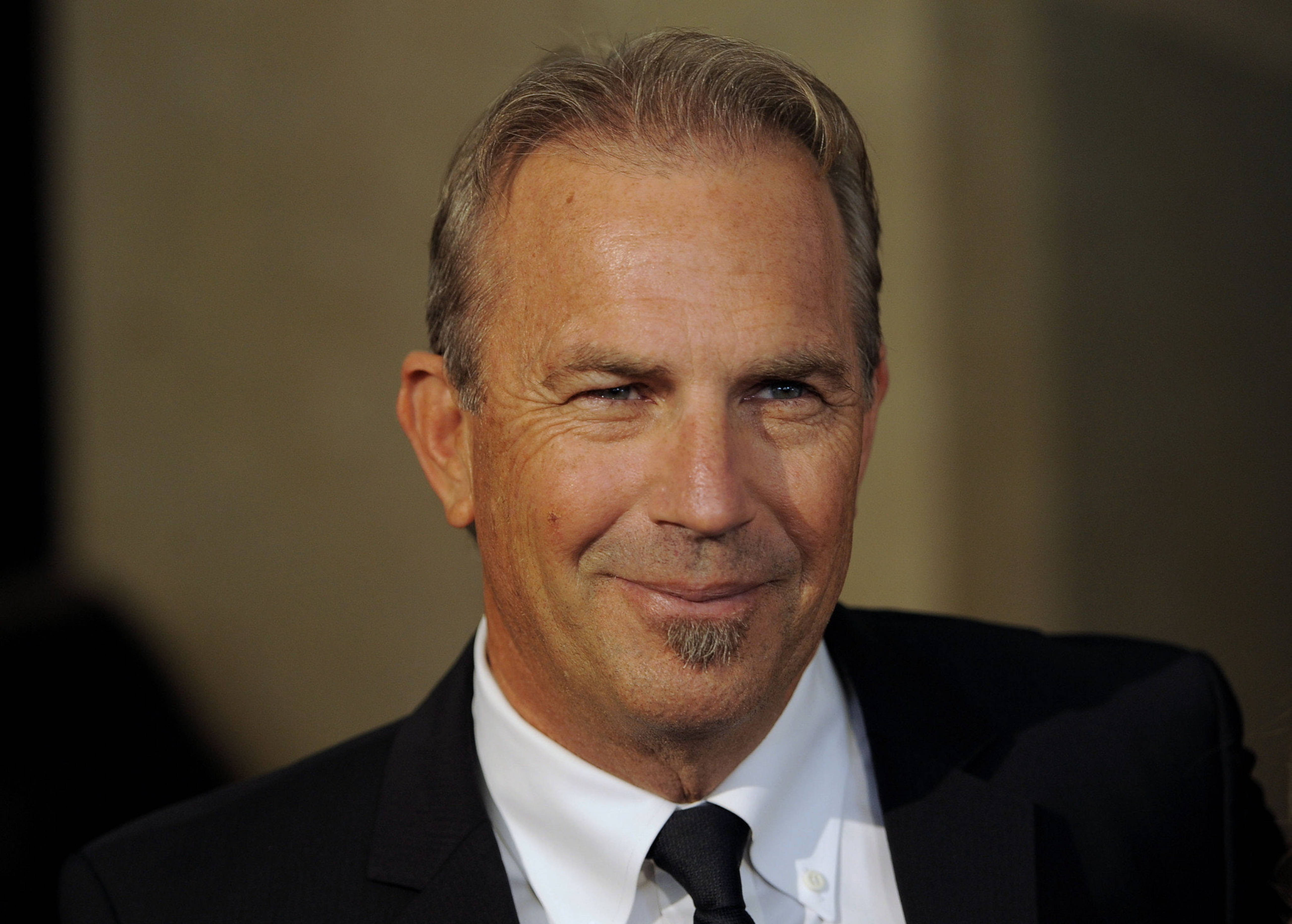 Kevin Costner 桌布 壁紙 — 一位微笑的中年男子特寫肖像，灰髮和山羊鬍鬚，穿著黑色西裝和領帶。