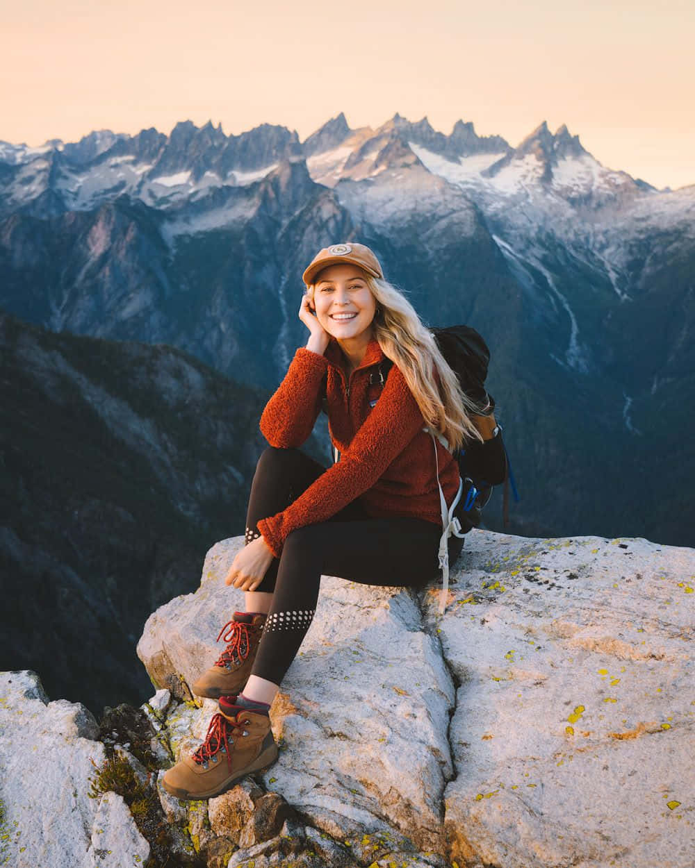 Winter Hiking 桌布 壁紙 — 一位微笑的健行者穿著紅色刷毛衣坐在岩石山頂上，身後是黃金時刻的雪峰。
