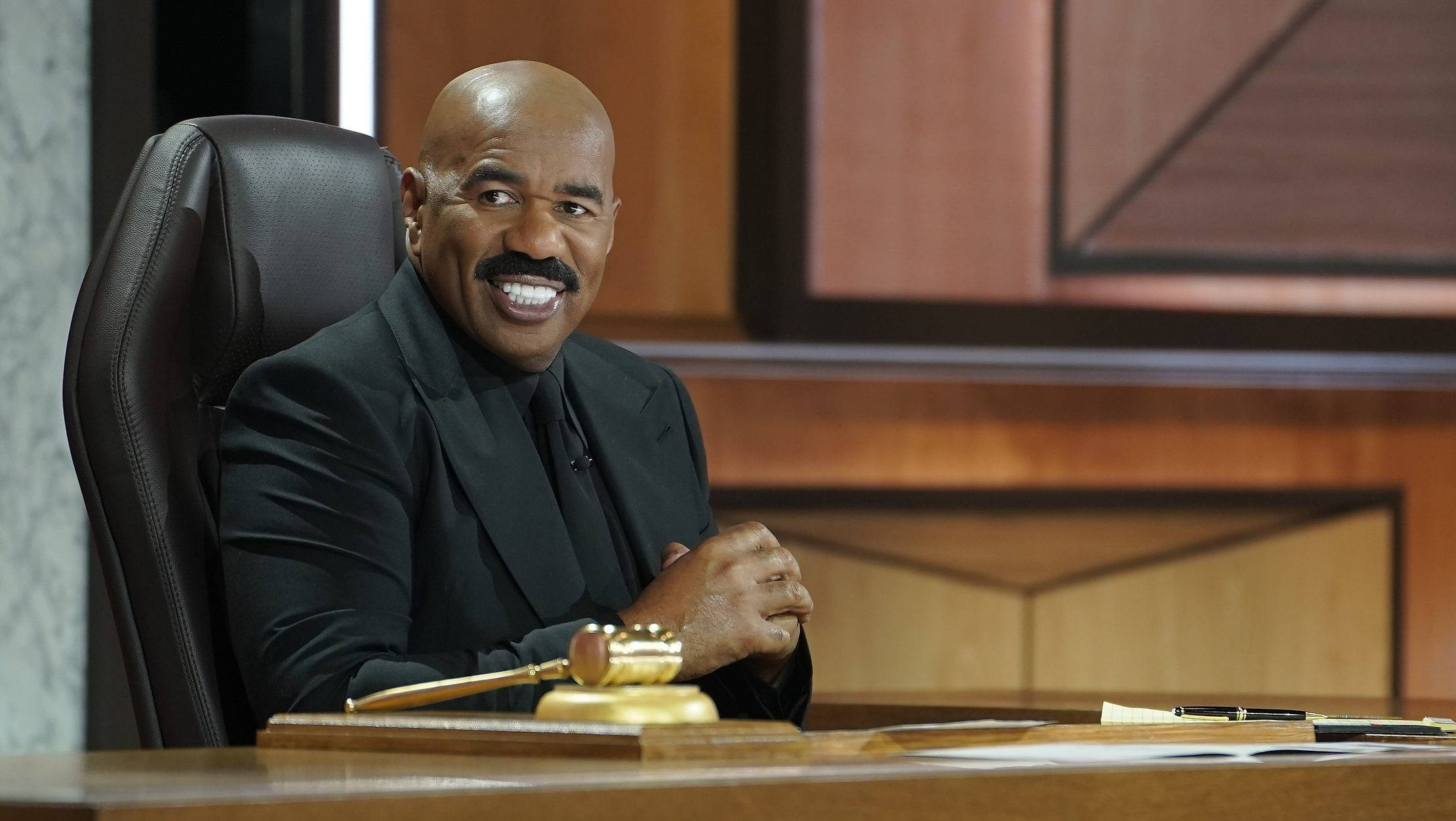steve harvey 桌布 壁紙 — 一位微笑的光頭男士留著鬍子，自信地坐在皮革椅上，身後是木製辦公桌，桌上放著木槌。