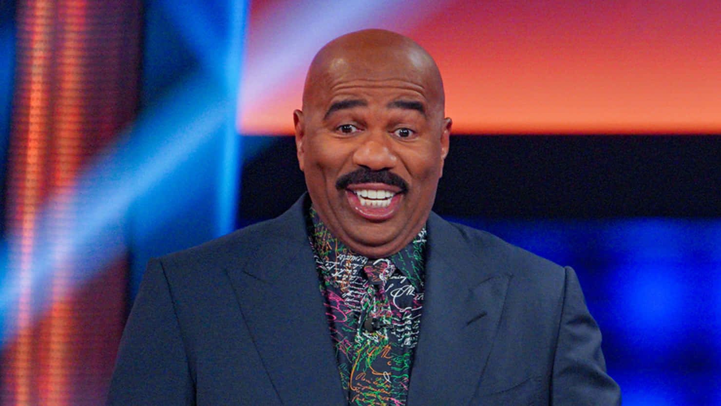 steve harvey 桌布 壁紙 — 一位微笑的光頭男士留著鬍子，身穿深色西裝和彩色圖案襯衫，背景是充滿活力的藍色和橙色工作室背景。
