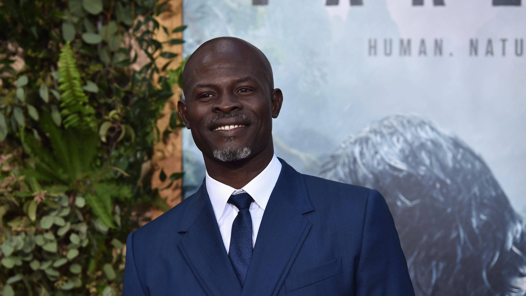 Celebrity 桌布 壁紙 — 一位微笑的光頭男士穿著剪裁俐落的海軍藍西裝，站在電影海報前，身旁有茂盛的綠植。