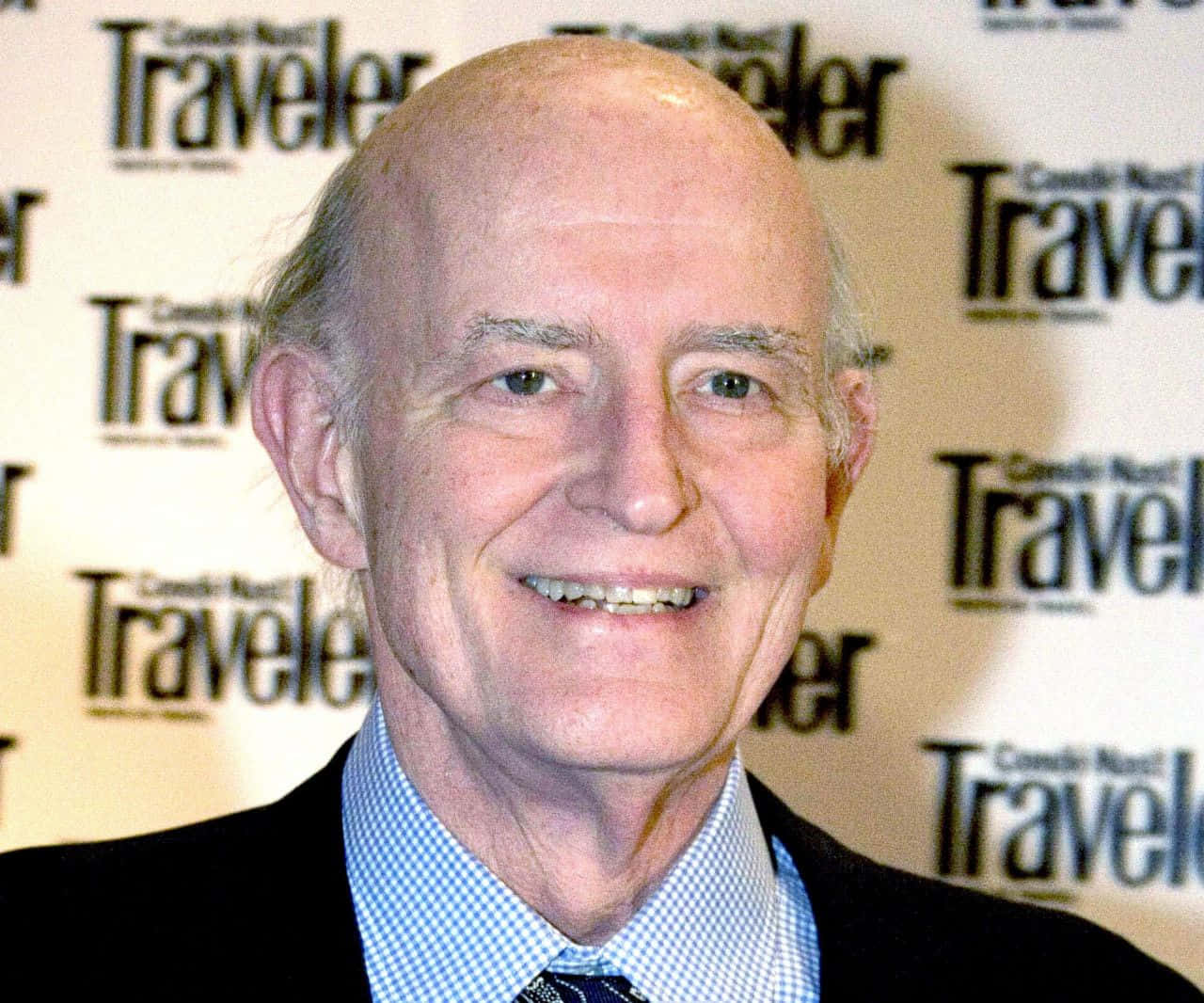 Peter Boyle 桌布 壁紙 — 一位微笑的光頭男士穿著西裝打領帶，站在重複的Traveler雜誌背景前。
