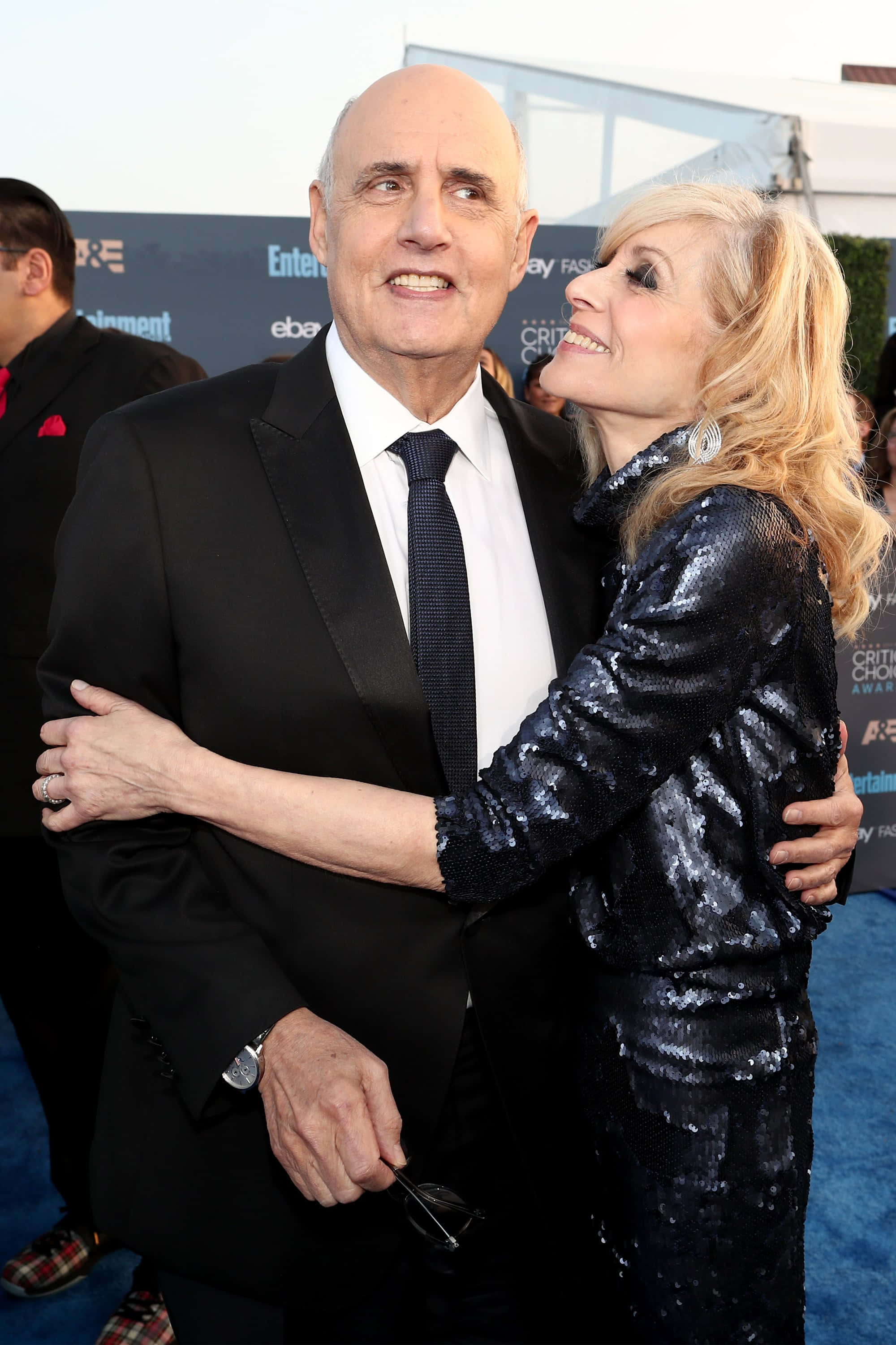 jeffrey tambor 桌布 壁紙 — 一位微笑的光頭男士身穿黑色西裝，在頒獎典禮的藍色地毯上擁抱一位身穿閃亮黑色連衣裙的金髮女士。