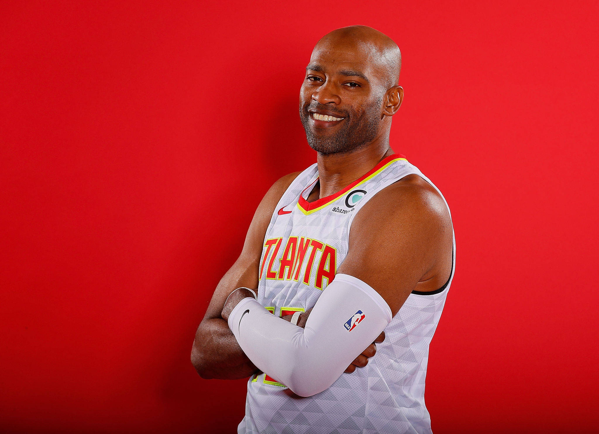 NBA 桌布 壁紙 — 一位微笑的光頭籃球運動員穿著亞特蘭大球衣，雙臂交叉，自信地站在純紅色背景前。