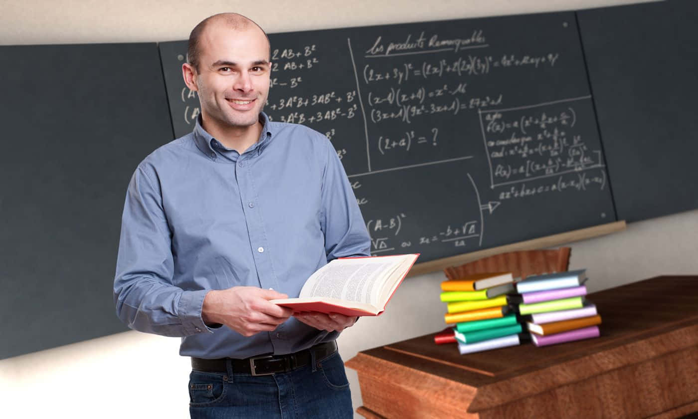 teachers 桌布 壁紙 — 一位微笑的光頭老師穿著藍色襯衫，在充滿數學公式的黑板和木桌上色彩繽紛的堆疊書籍前，拿著一本紅色的書。
