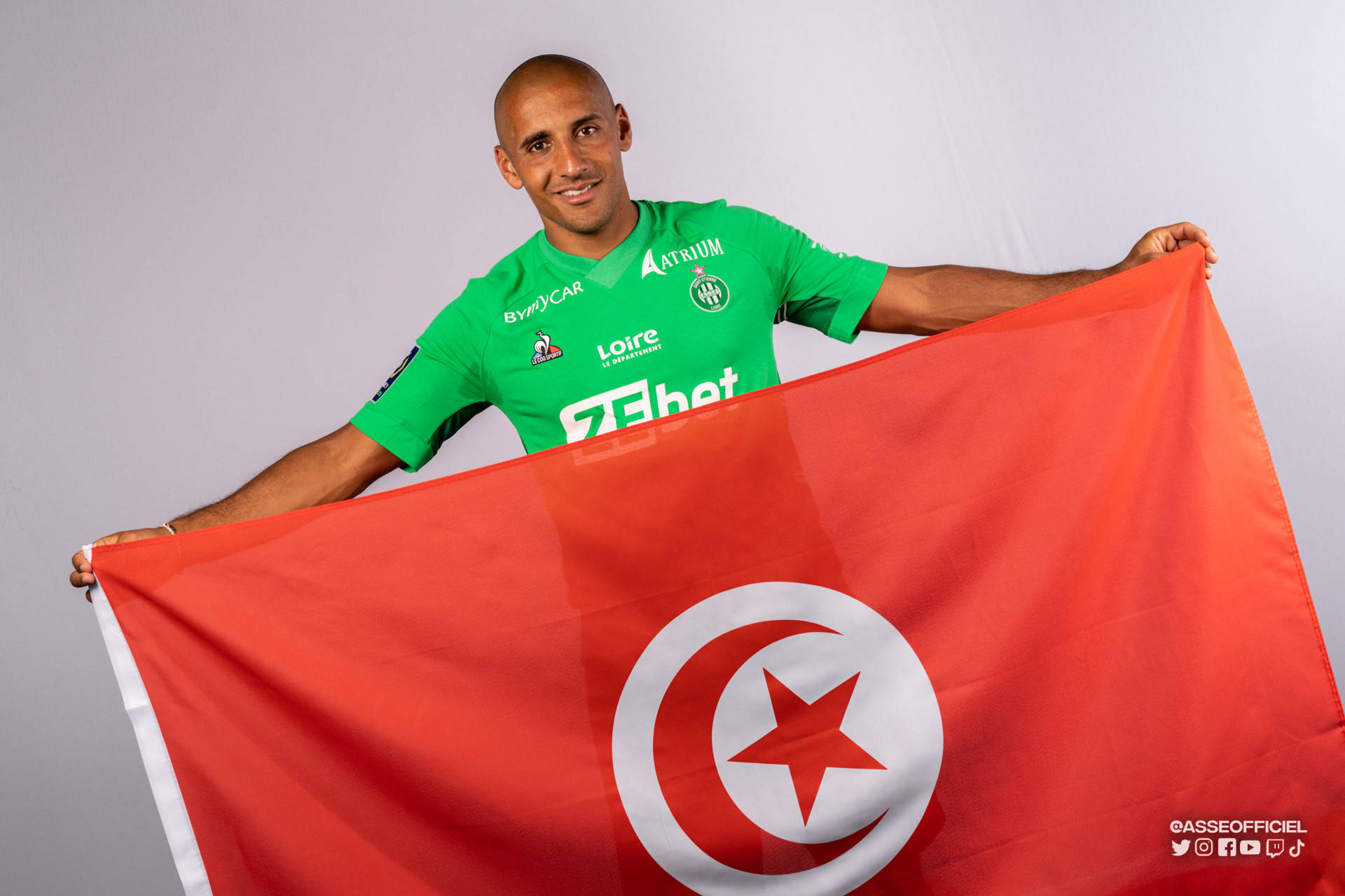 Tunisia 桌布 壁紙 — 一位微笑的光頭足球員身穿綠色球衣，在純灰色攝影棚背景前自豪地舉起一面帶有白色新月和星星的大型紅色突尼斯國旗。