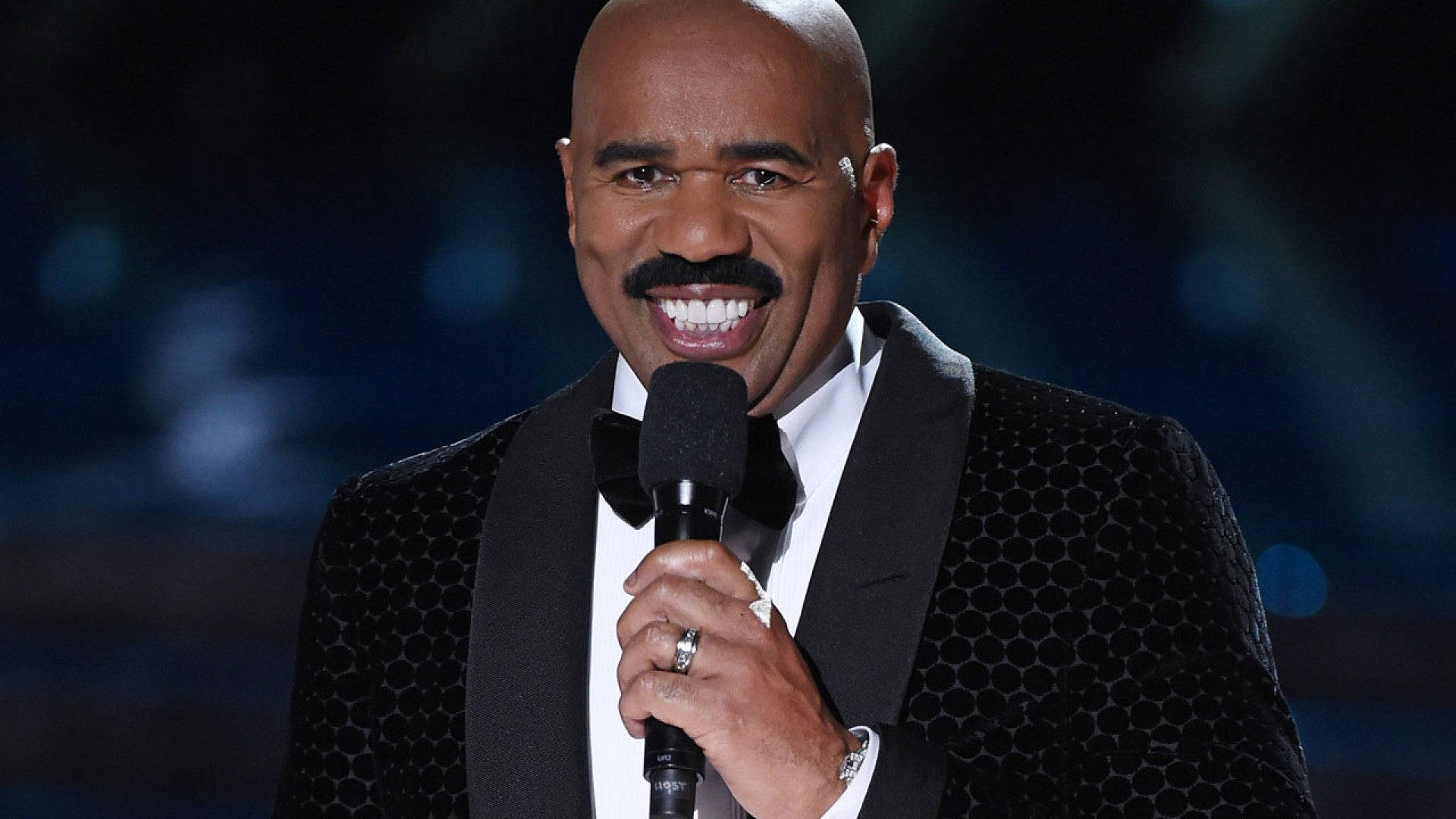 steve harvey 桌布 壁紙 — 一位微笑的光頭鬍鬚男，身穿黑色圖案禮服，在藍色燈光的黑暗舞台上手持麥克風。