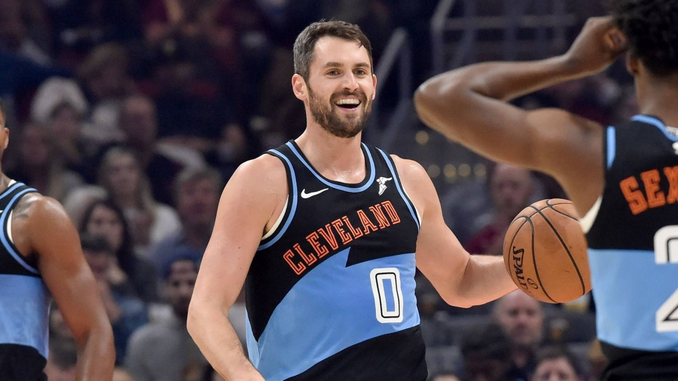 Kevin Love 桌布 壁紙 — 一位微笑的克里夫蘭籃球運動員身穿藍黑球衣，在激烈的比賽中手持籃球，背景有隊友和觀眾。