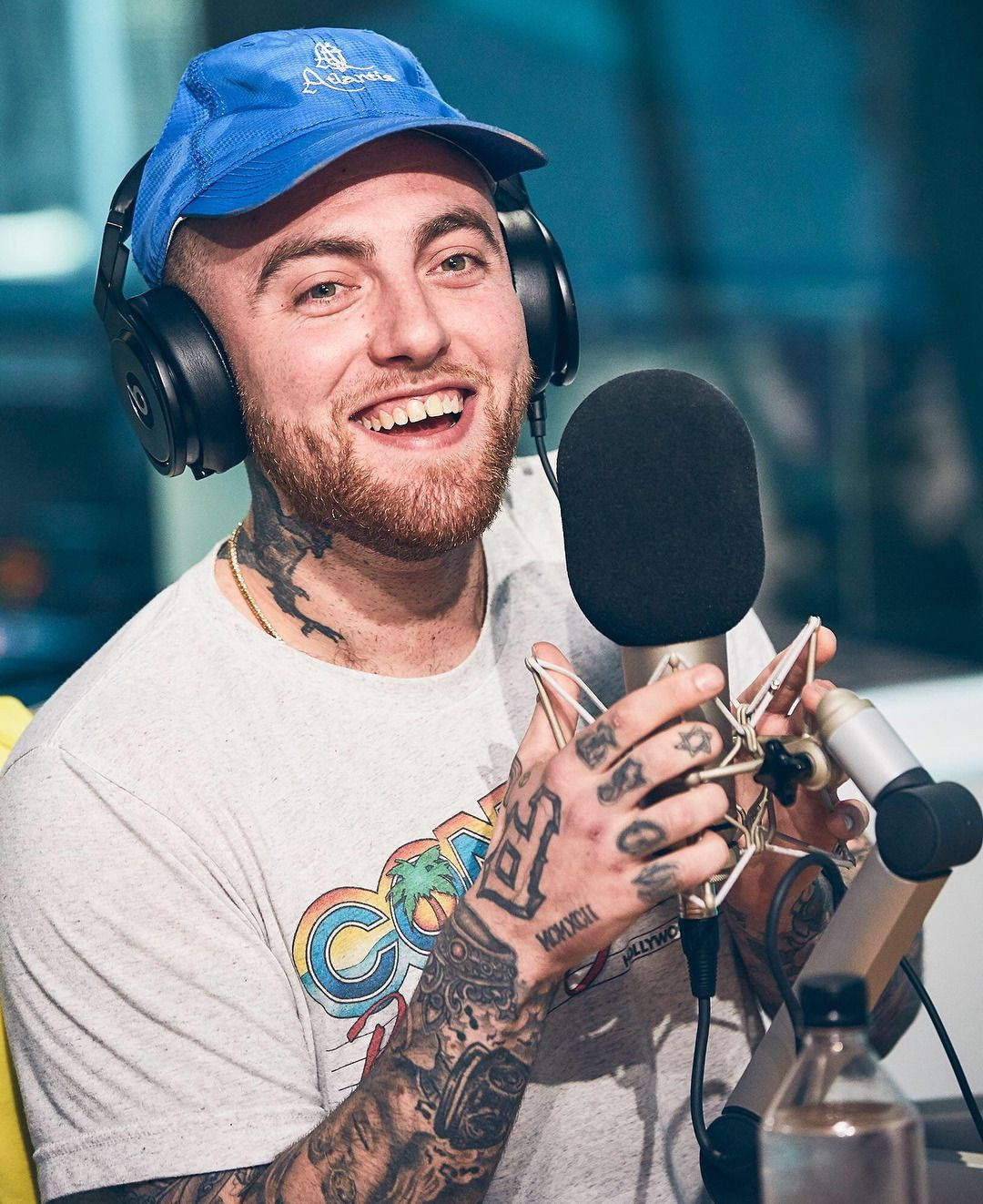 Mac Miller 桌布 壁紙 — 一位微笑的刺青男子戴著藍色帽子和耳機，在錄音室裡拿著麥克風。
