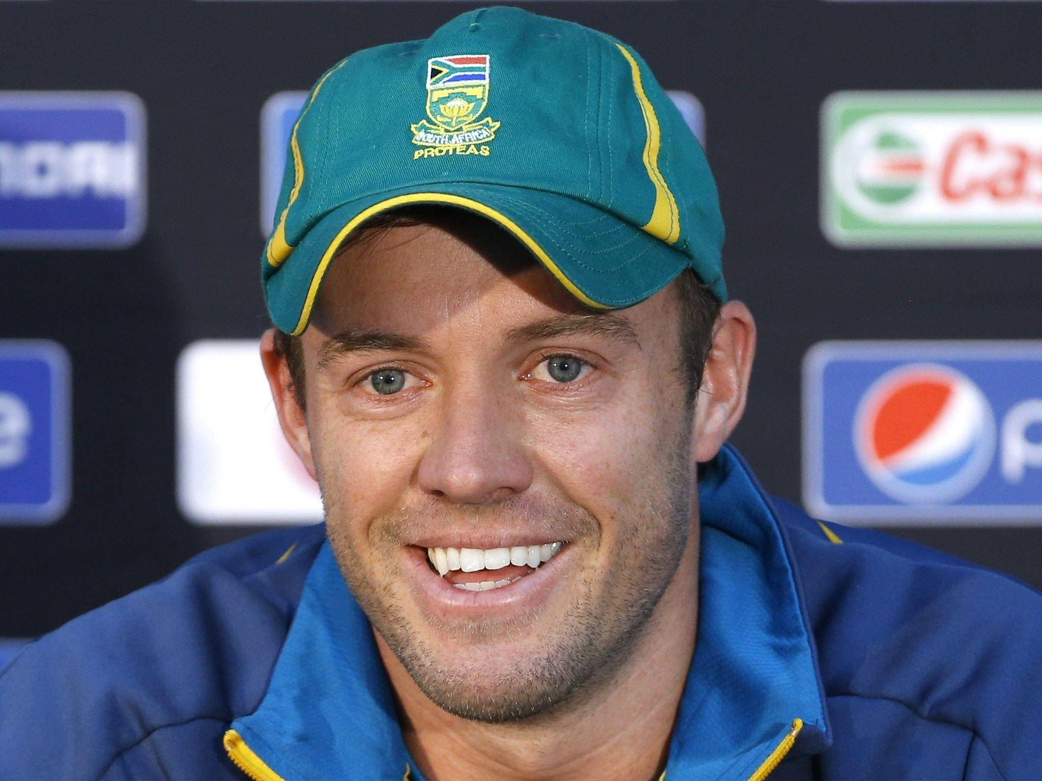 Ab De Villiers 桌布 壁紙 — 一位微笑的南非板球運動員，戴著綠色帽子、穿著藍色外套，在記者會上擺姿勢，身後有贊助商標誌。