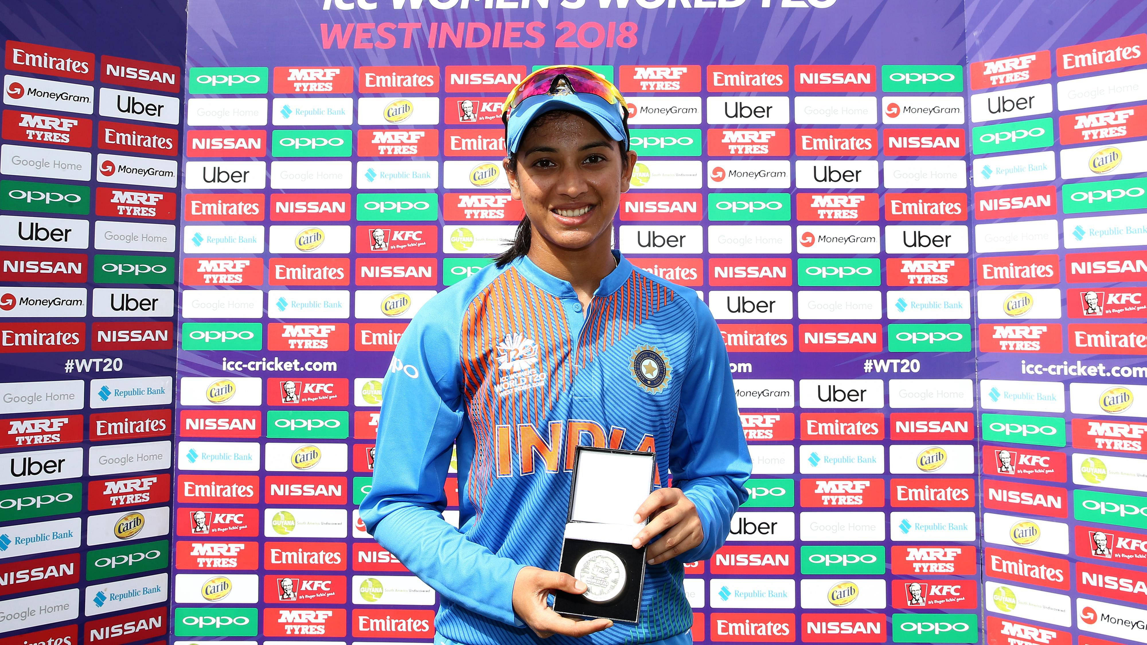 Smriti Mandhana 桌布 壁紙 — 一位微笑的印度女子板球運動員身穿藍色制服，在2018年ICC女子世界T20賽事中，於色彩繽紛的贊助商牆前手持銀牌。