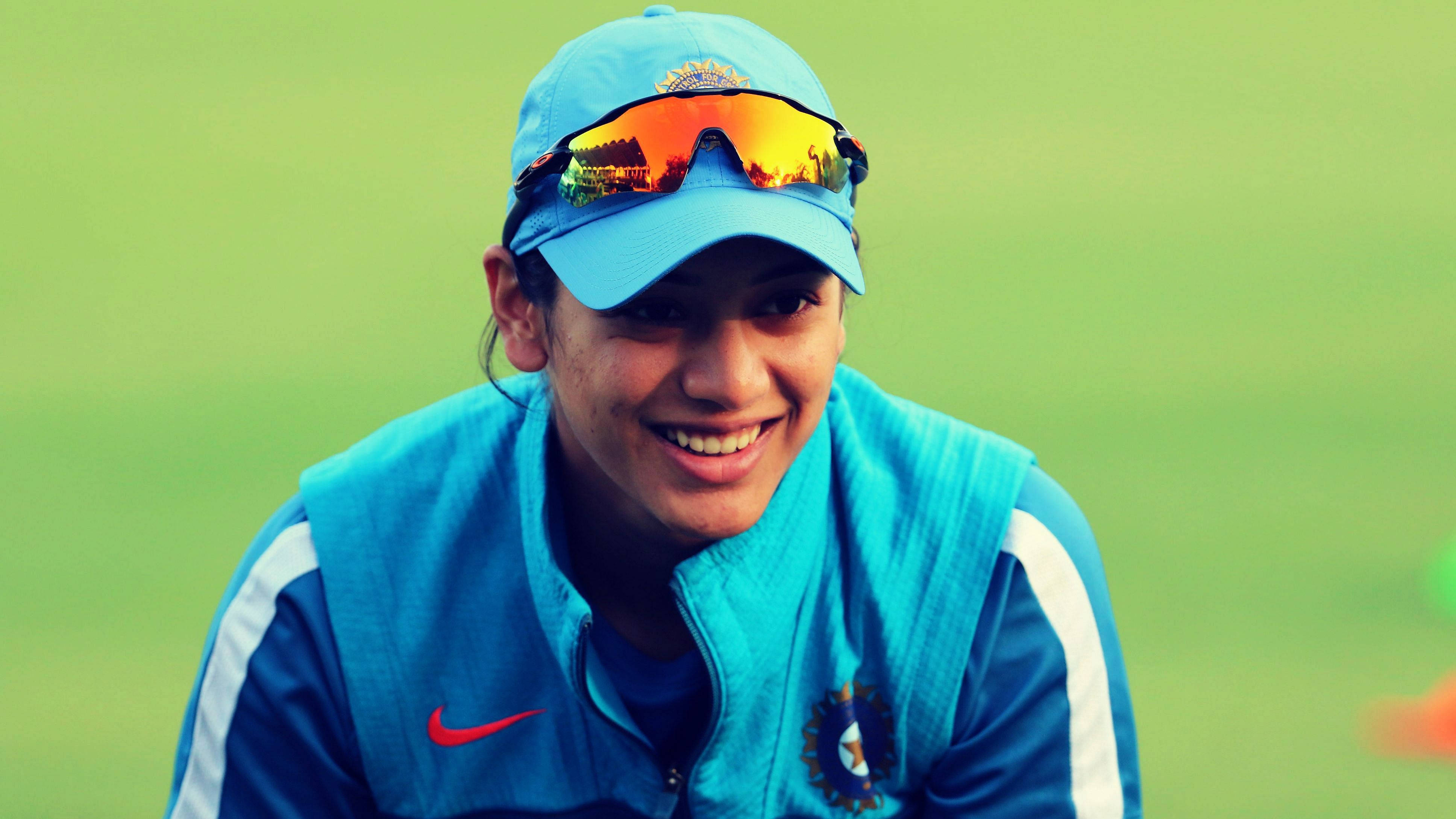Smriti Mandhana 桌布 壁紙 — 一位微笑的印度板球運動員，身穿藍色裝備和帽子，頭戴反光太陽眼鏡，背景是綠色球場。