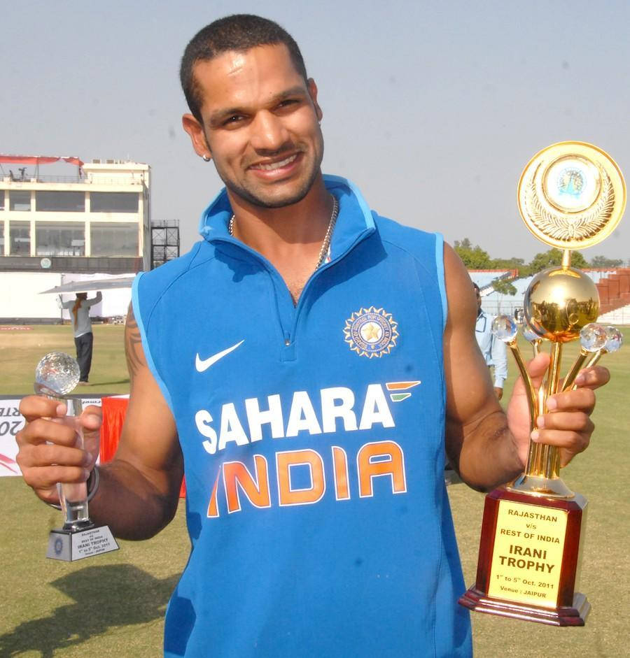 shikhar dhawan 桌布 壁紙 — 一位微笑的印度板球運動員身穿藍色Sahara India球衣，在陽光明媚的板球場上自豪地舉起兩個獎盃。
