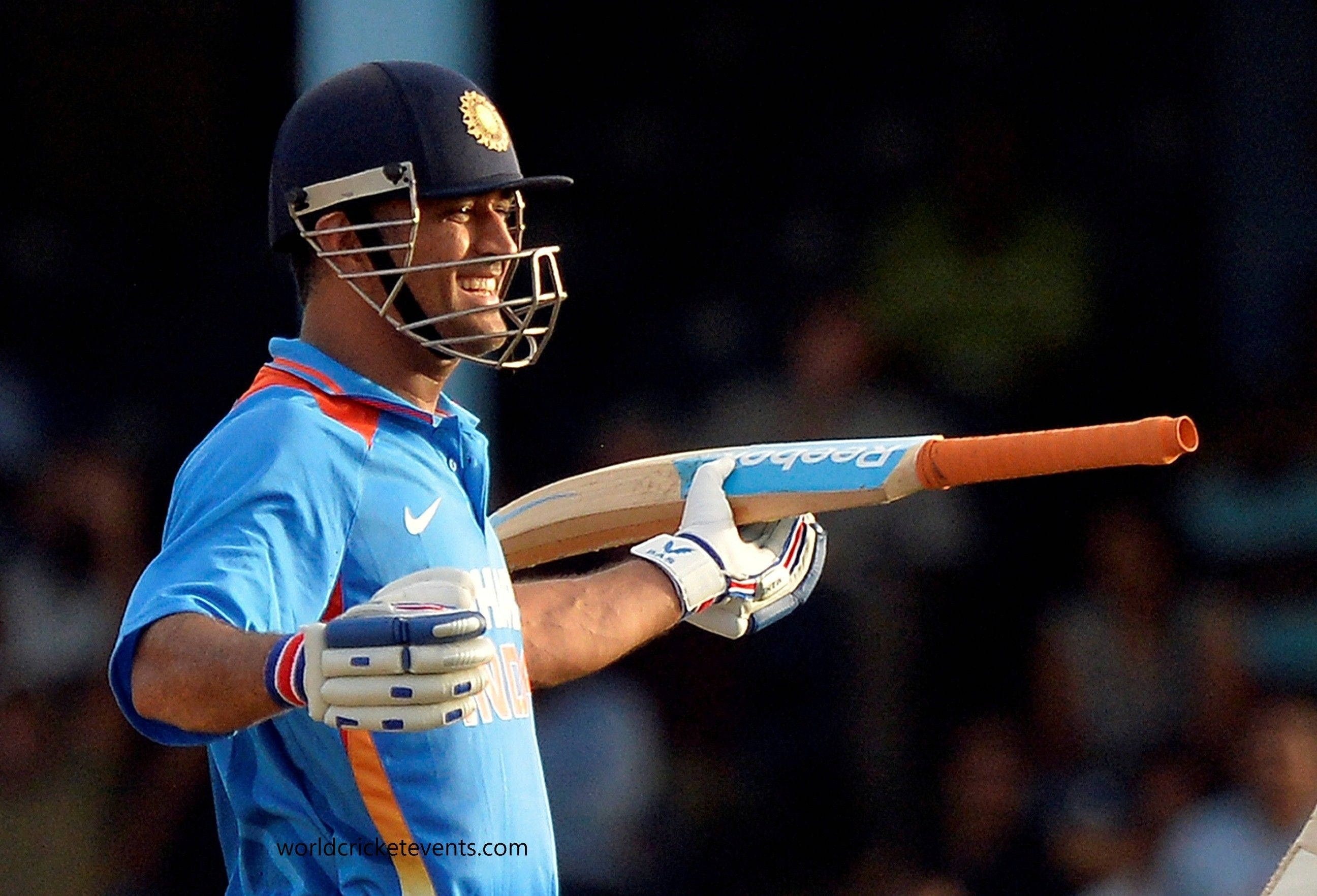 MS Dhoni 桌布 壁紙 — 一位微笑的印度板球運動員身穿藍色制服，在模糊的體育場人群背景下將球棒扛在肩上。