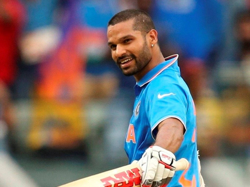 shikhar dhawan 桌布 壁紙 — 一位微笑的印度板球運動員身穿藍色球衣，在比賽中手持球棒，背景是模糊的體育場人群。