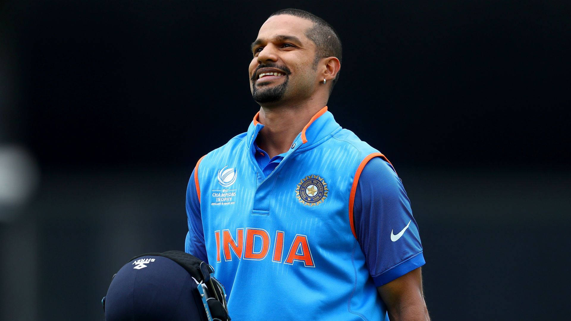 shikhar dhawan 桌布 壁紙 — 一位微笑的印度板球運動員身穿藍色訓練裝備，在深色背景下手持頭盔。