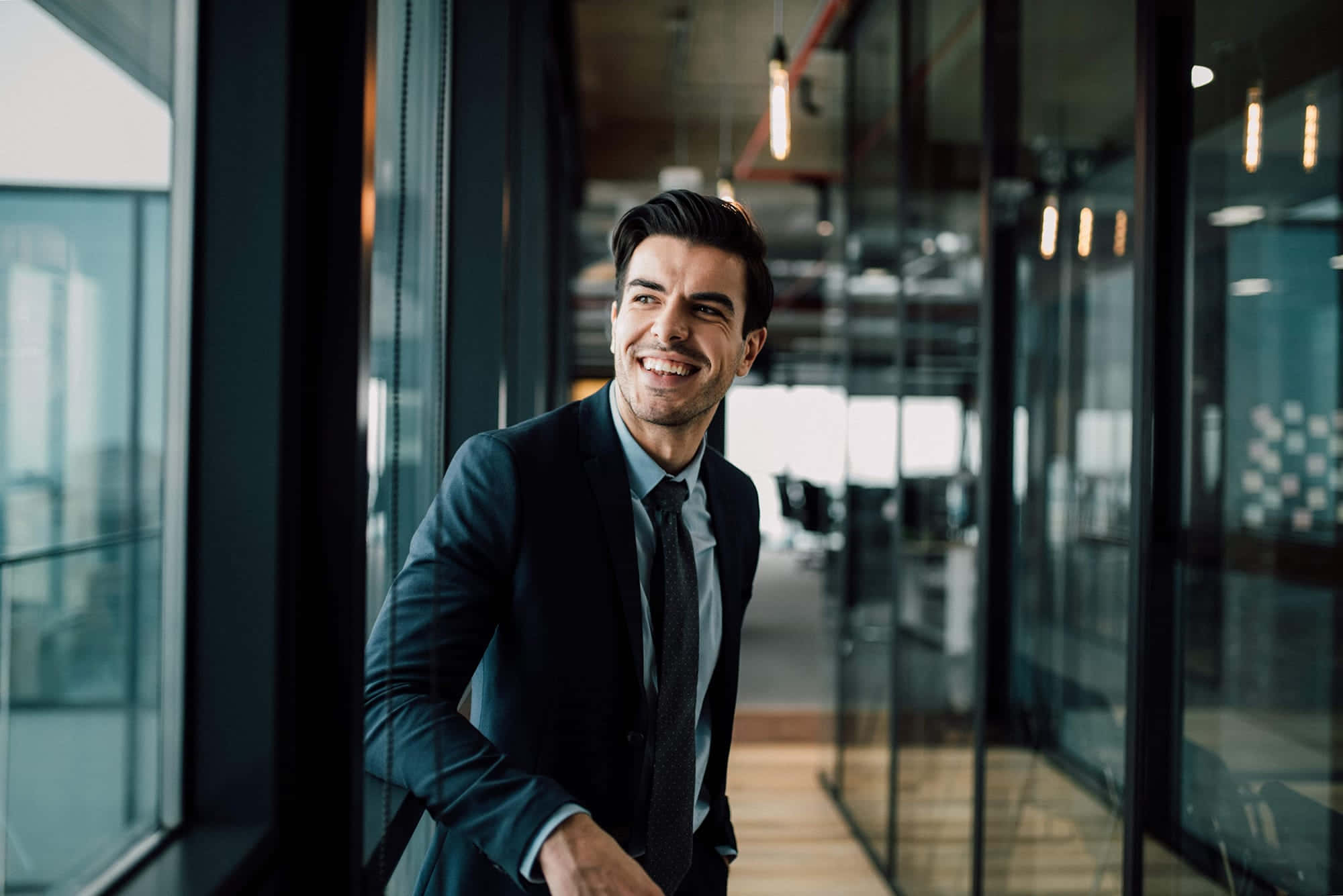 Real Estate Agent 桌布 壁紙 — 一位微笑的商人穿著西裝，靠在現代照明背景的玻璃辦公室門上。