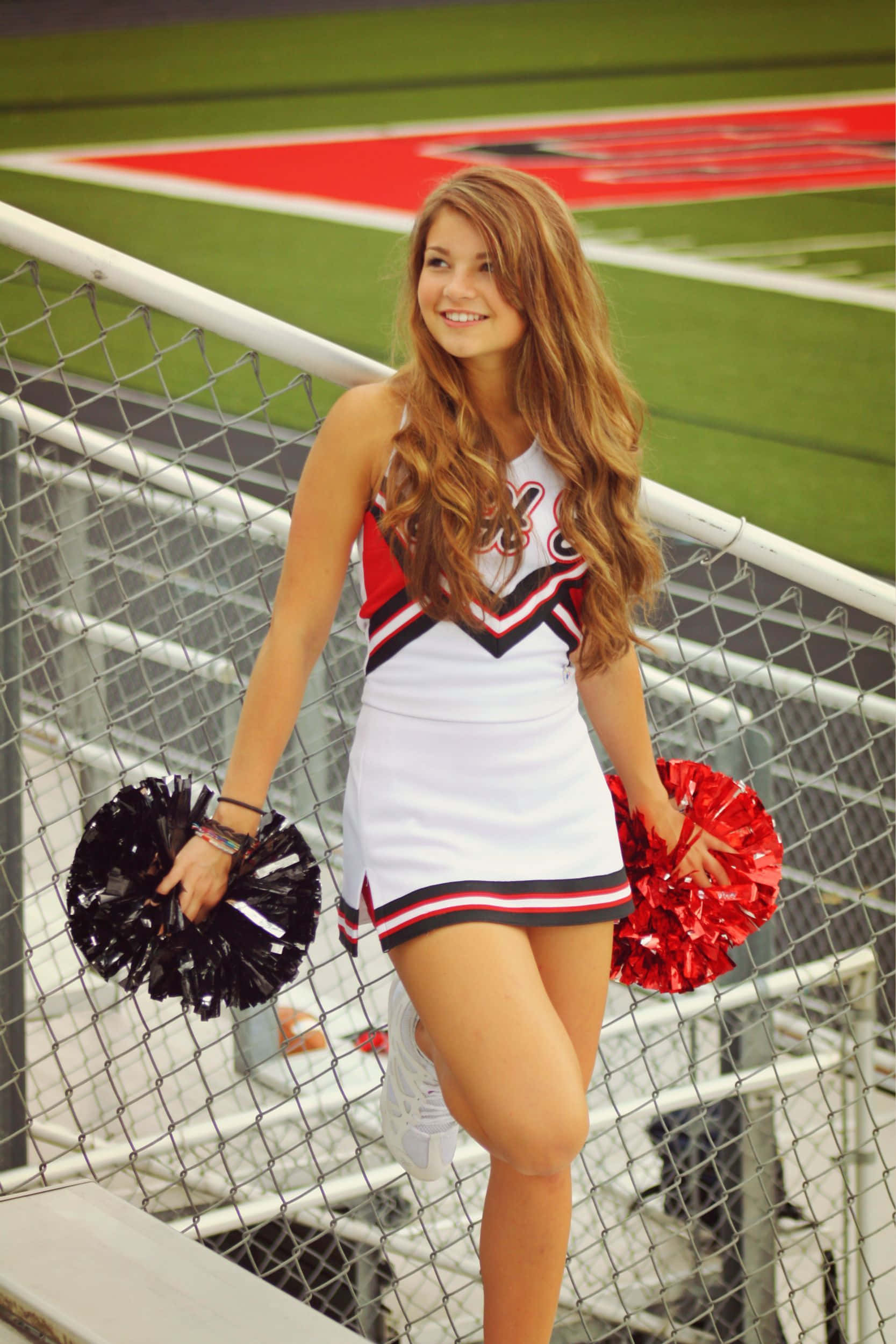 Cheer 桌布 壁紙 — 一位微笑的啦啦隊員身穿白色、紅色和黑色制服，在足球場圍欄旁拿著彩球擺姿勢。
